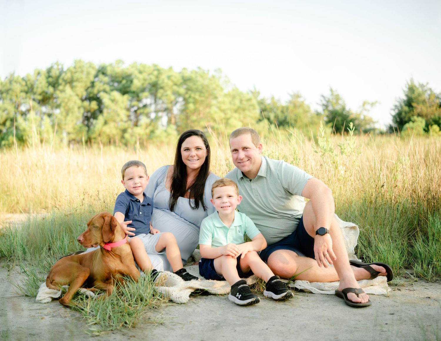 A family sitting together on a blanket in tall golden grass with their brown dog during an outdoor photo session.