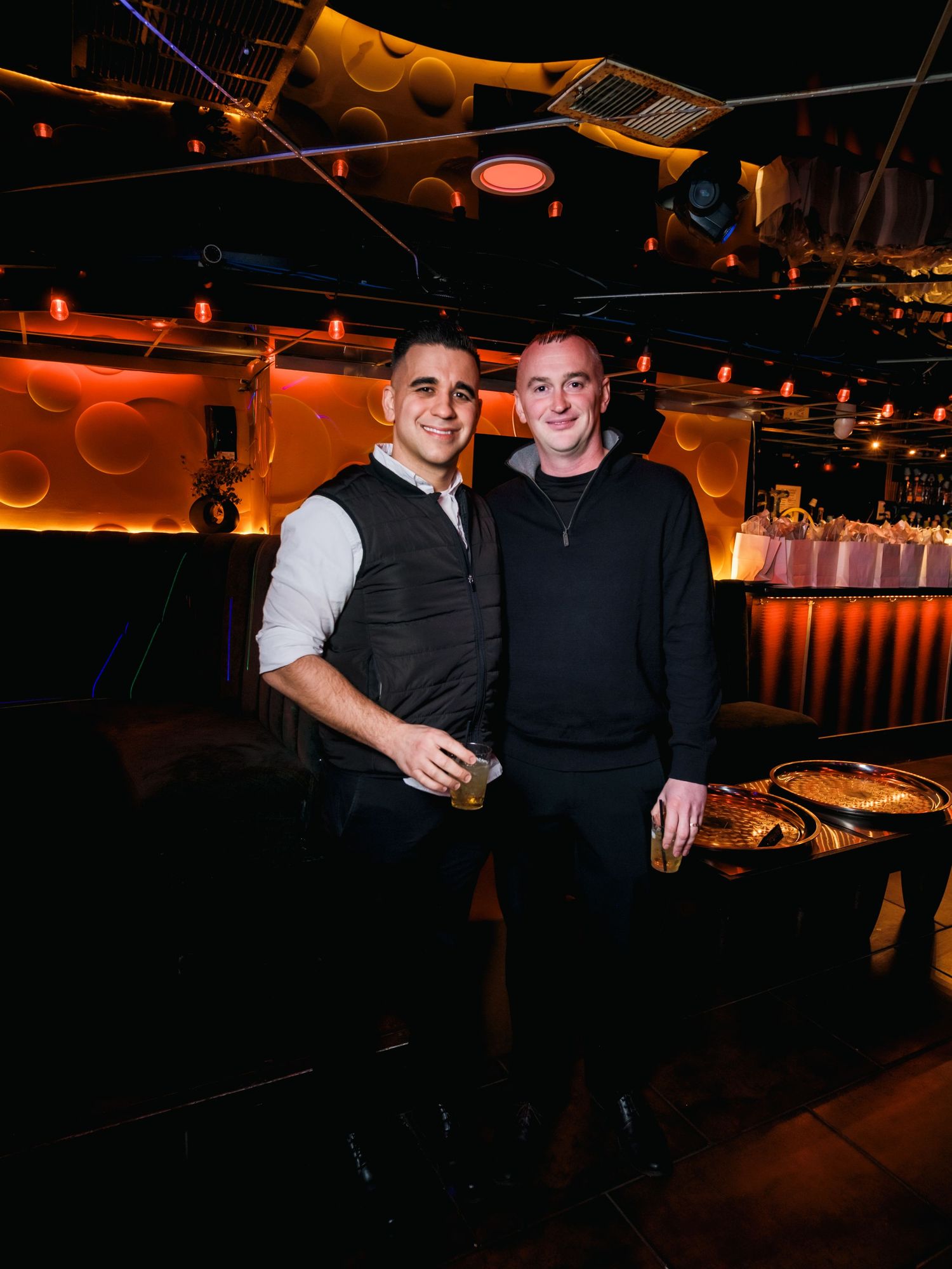 Two friends pose together inside a dimly lit lounge with warm lighting and decorative elements in background.