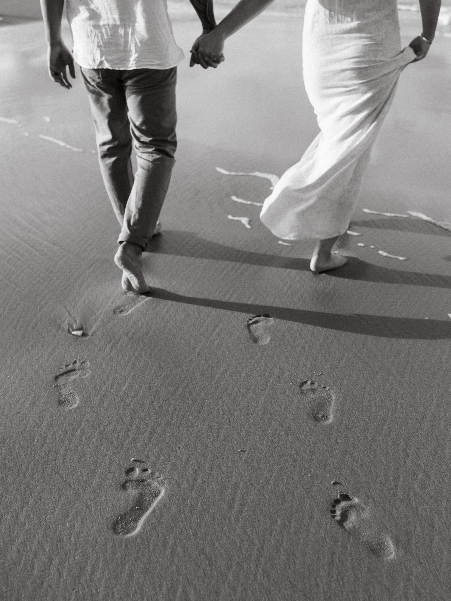 Footprints in wet sand lead towards the shore in black and white.