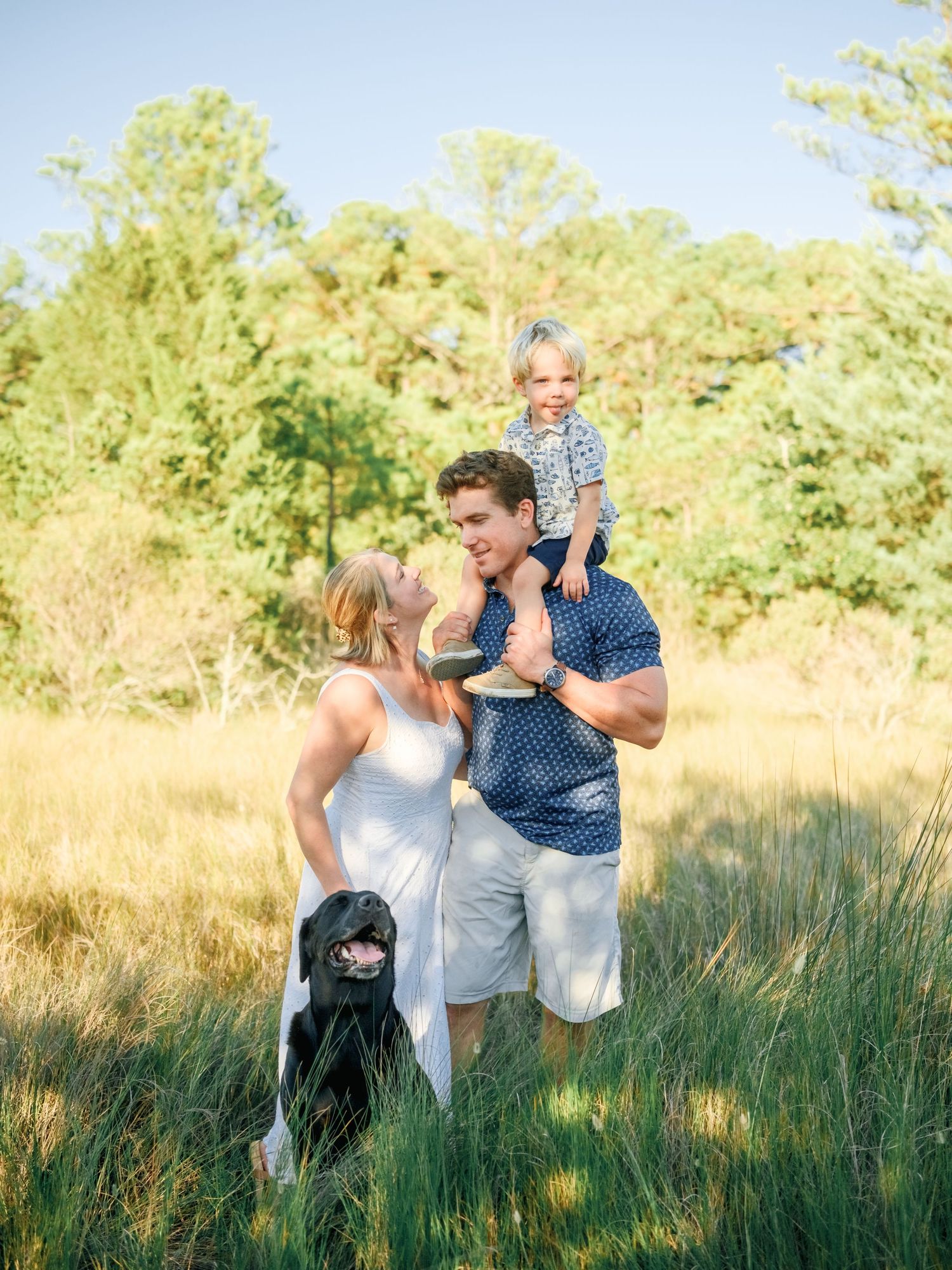 A couple plays and laughs together in a grassy field with their black dog on a sunny afternoon.