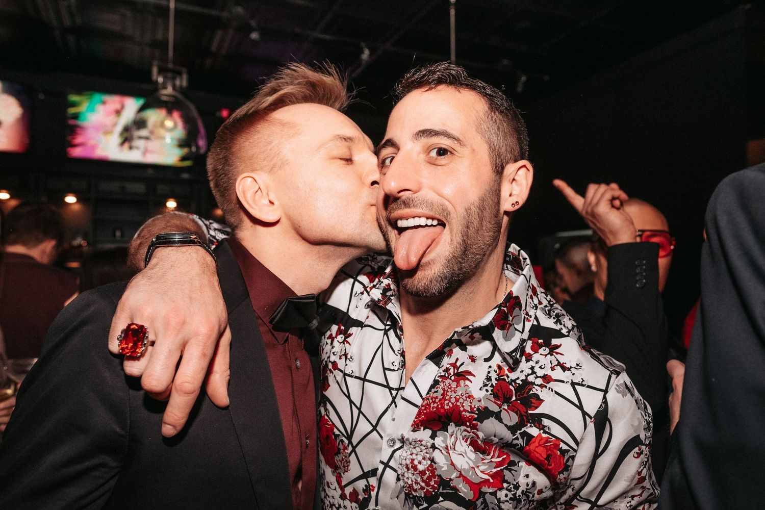 Two friends celebrating at a nightclub with one kissing the other's cheek while embracing in a dimly lit environment.