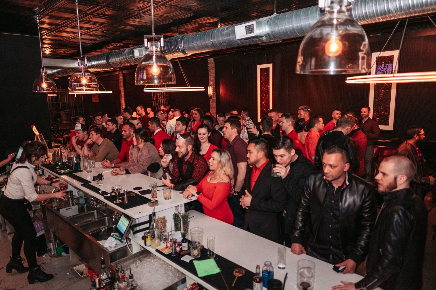 A packed nightclub crowd gathers in a dimly lit venue with exposed industrial ceiling and warm lighting.