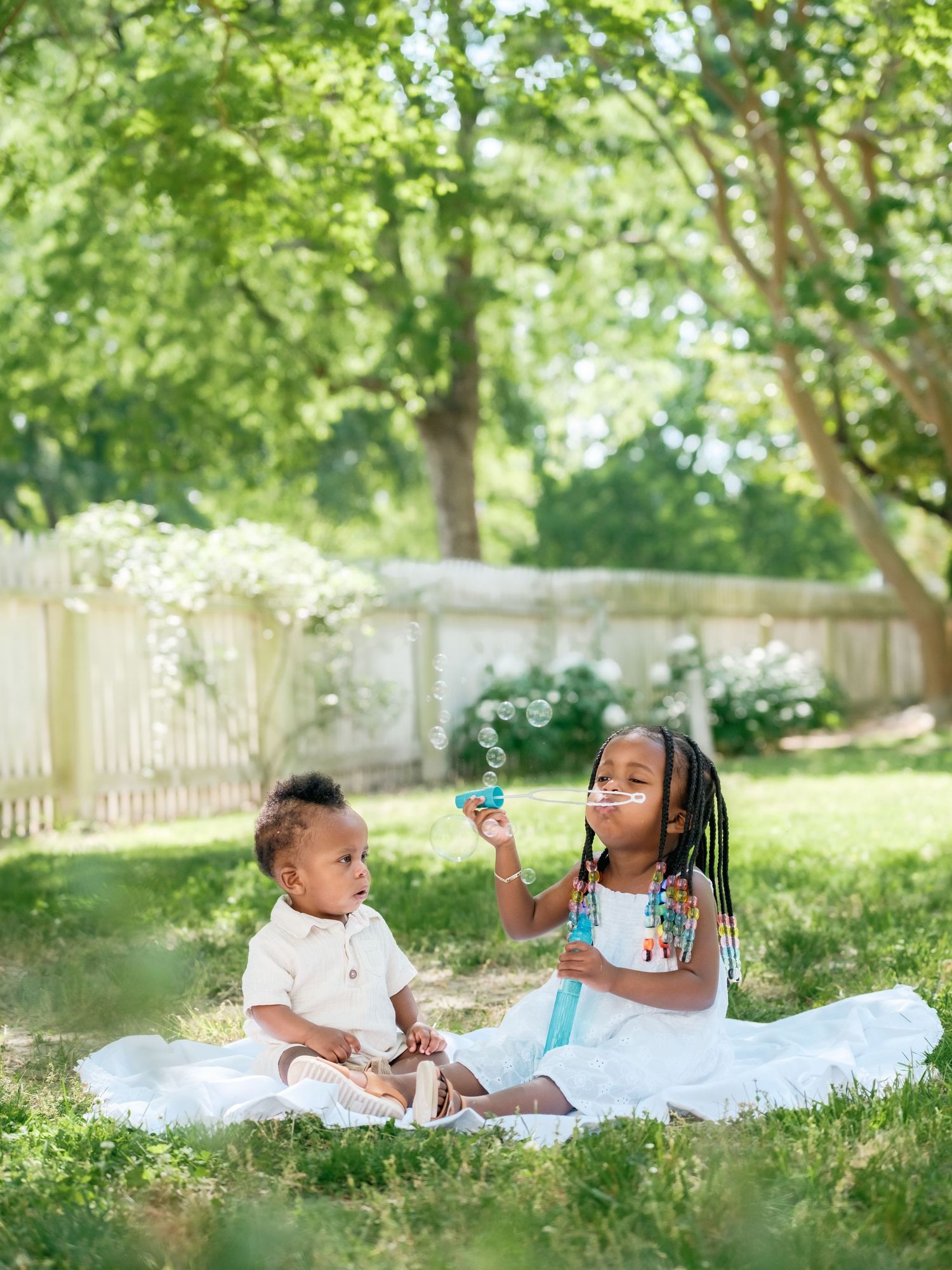 People enjoy a picnic on a white blanket under green trees during a sunny summer day in a lush garden setting.