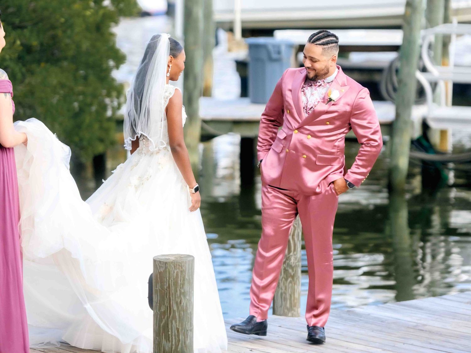 Wedding party members in white and pink attire pose together on a dock by the water on a sunny day.