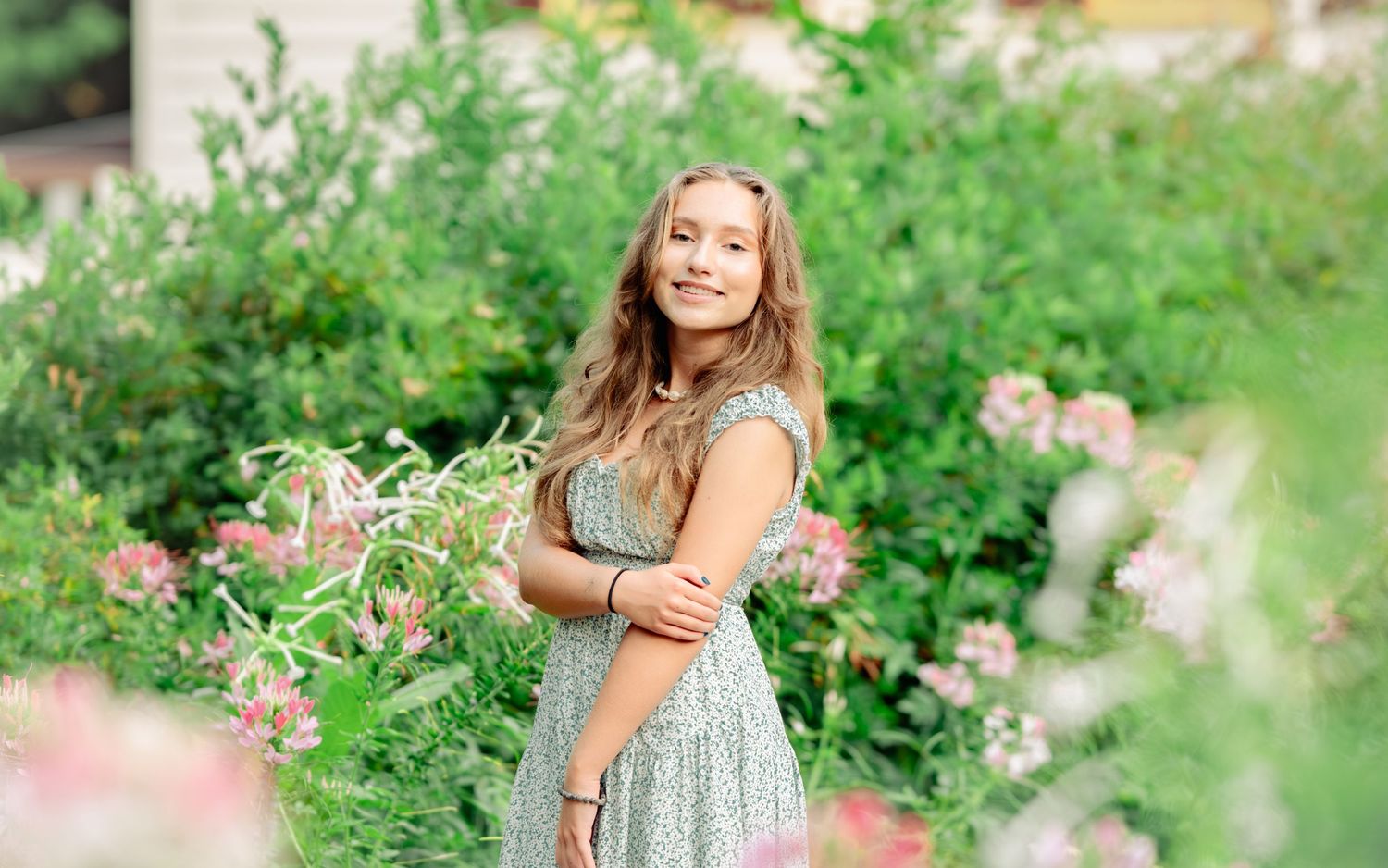 A cheerful young person in a floral dress sits surrounded by lush greenery and pink flowers in a garden setting.