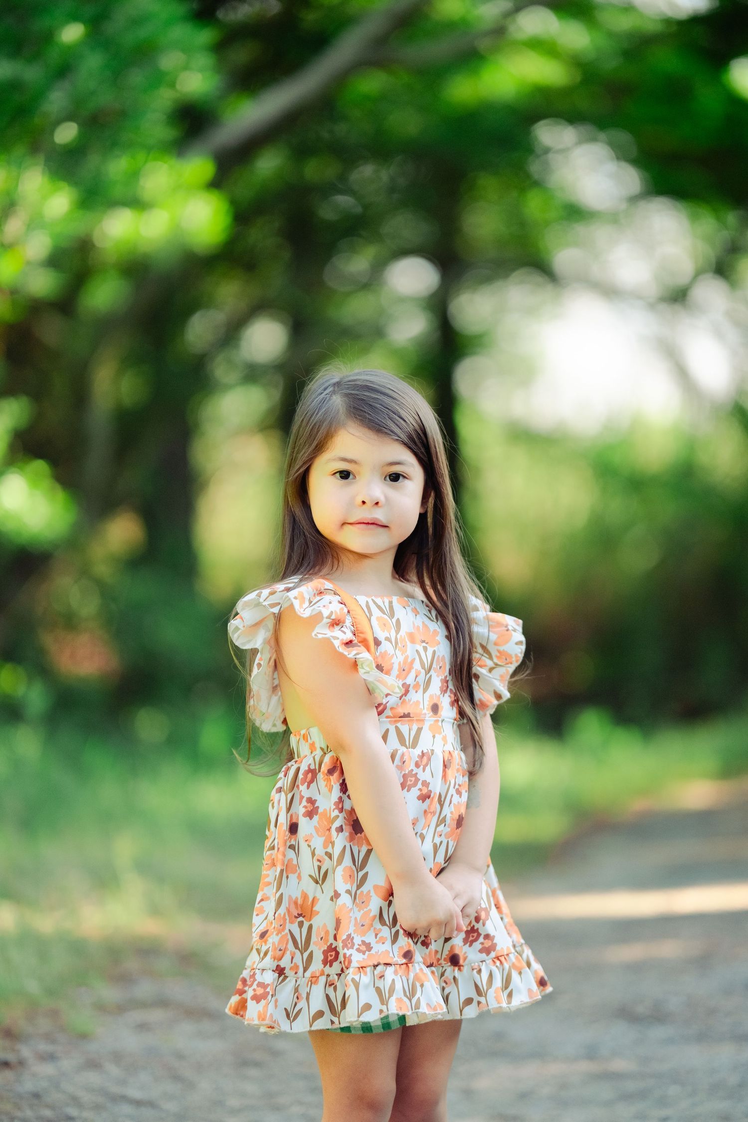 A child wearing a floral print dress with ruffled sleeves stands on a sunlit dirt path surrounded by green foliage.