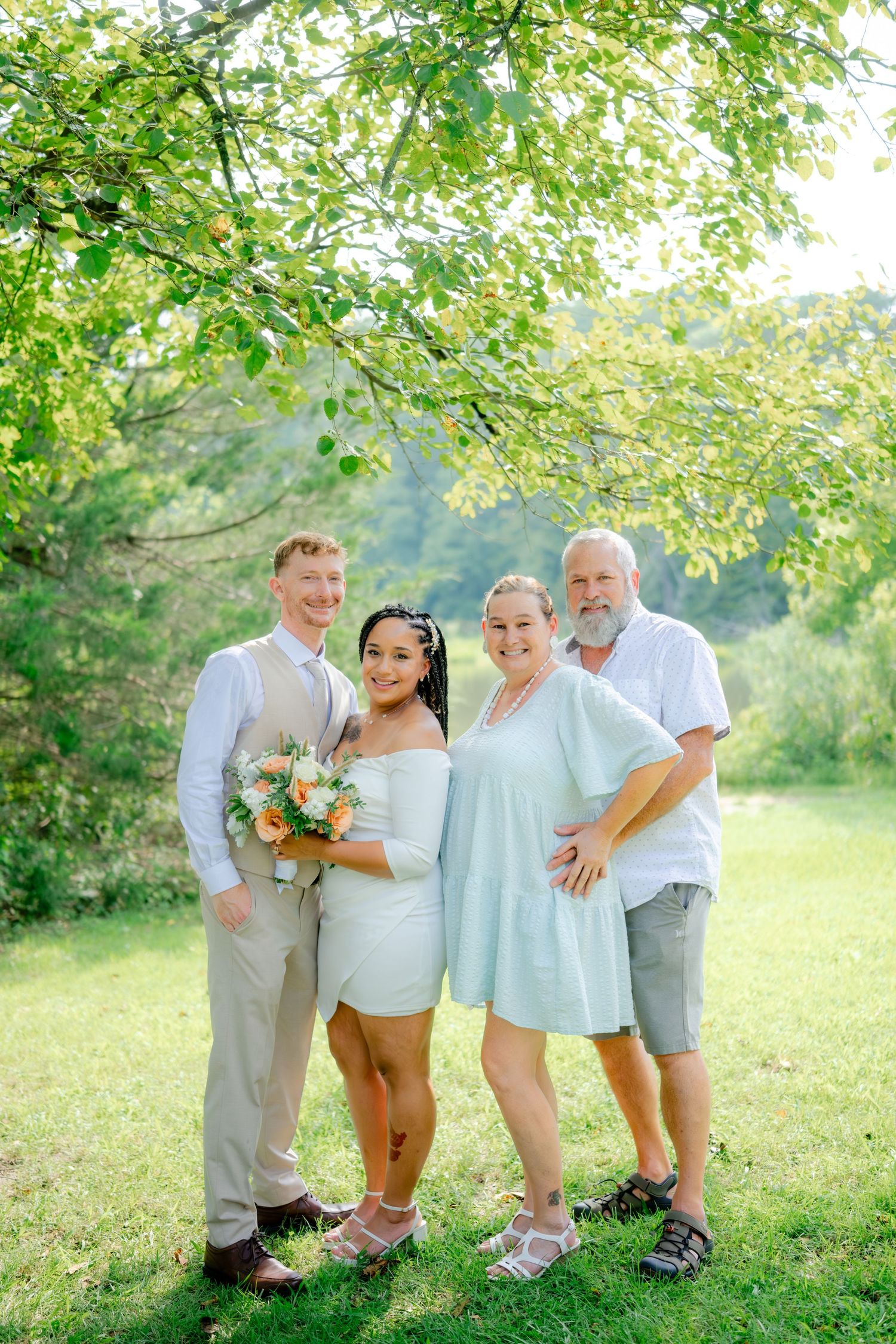 Four people stand together outdoors on a summer day beneath green trees while one holds a floral bouquet.
