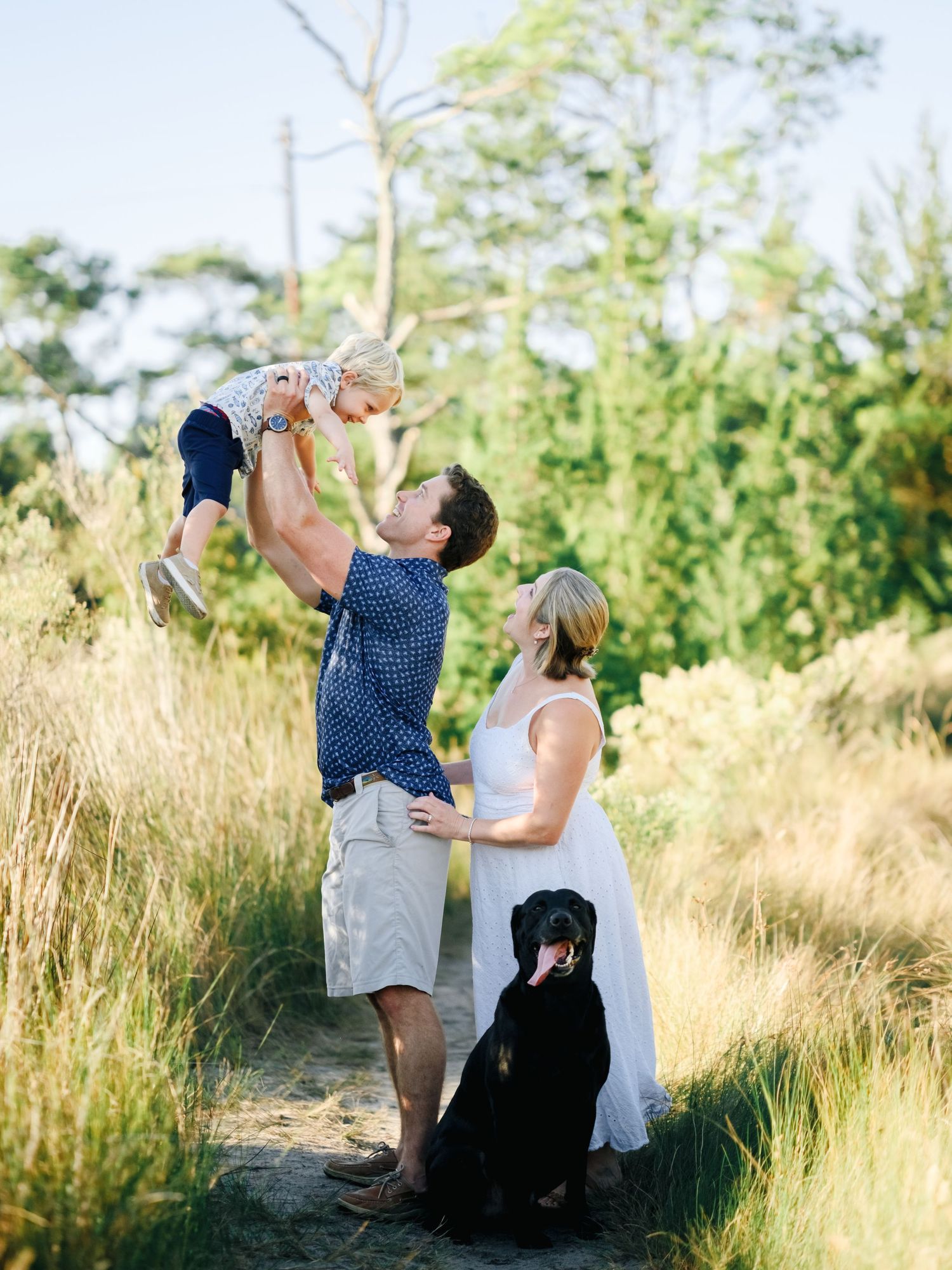 A couple shares romantic moments in a grassy field at sunset with their black dog beside them.