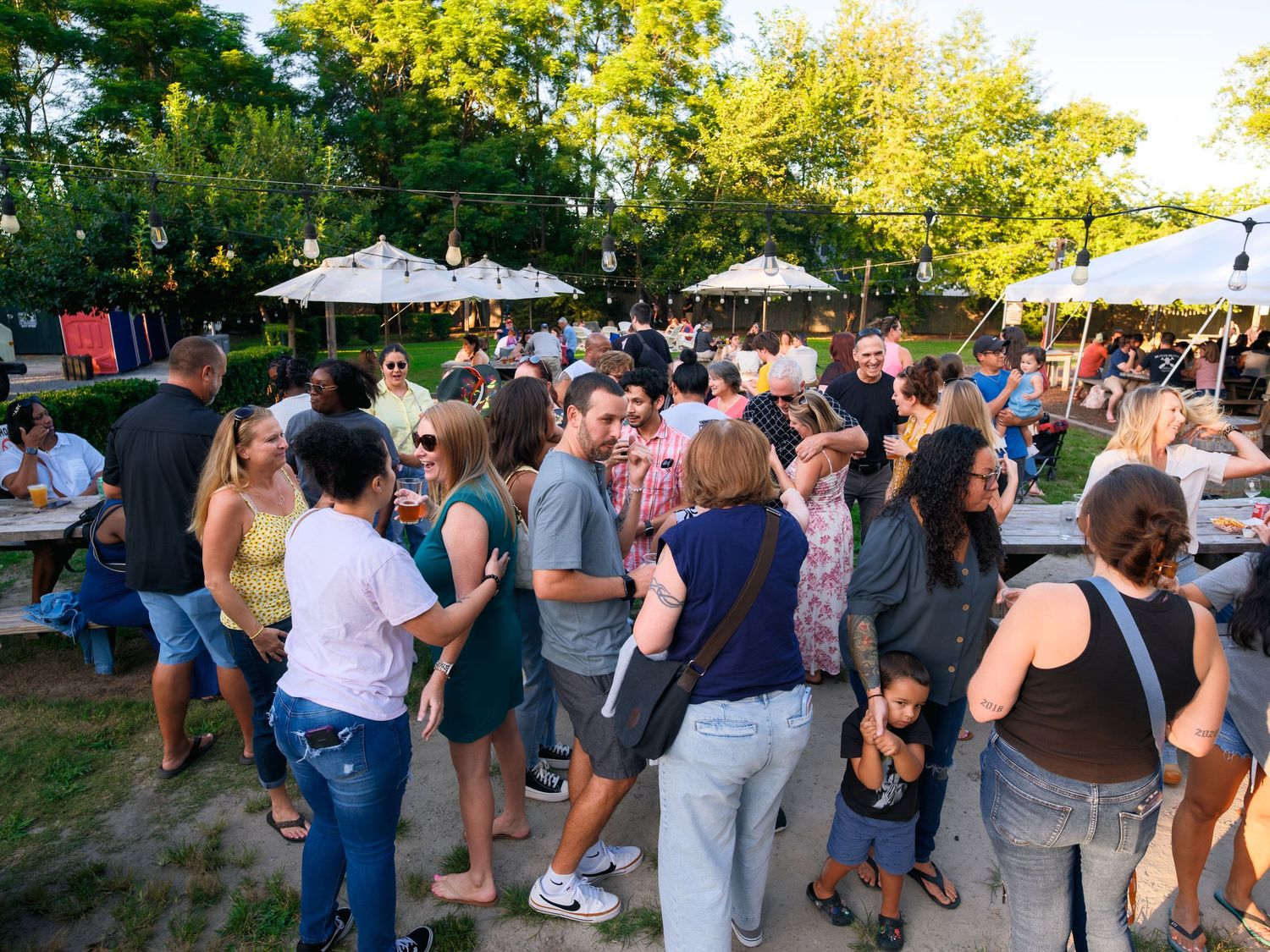 A crowd of people gathered at an outdoor summer event beneath shady trees and white umbrellas.