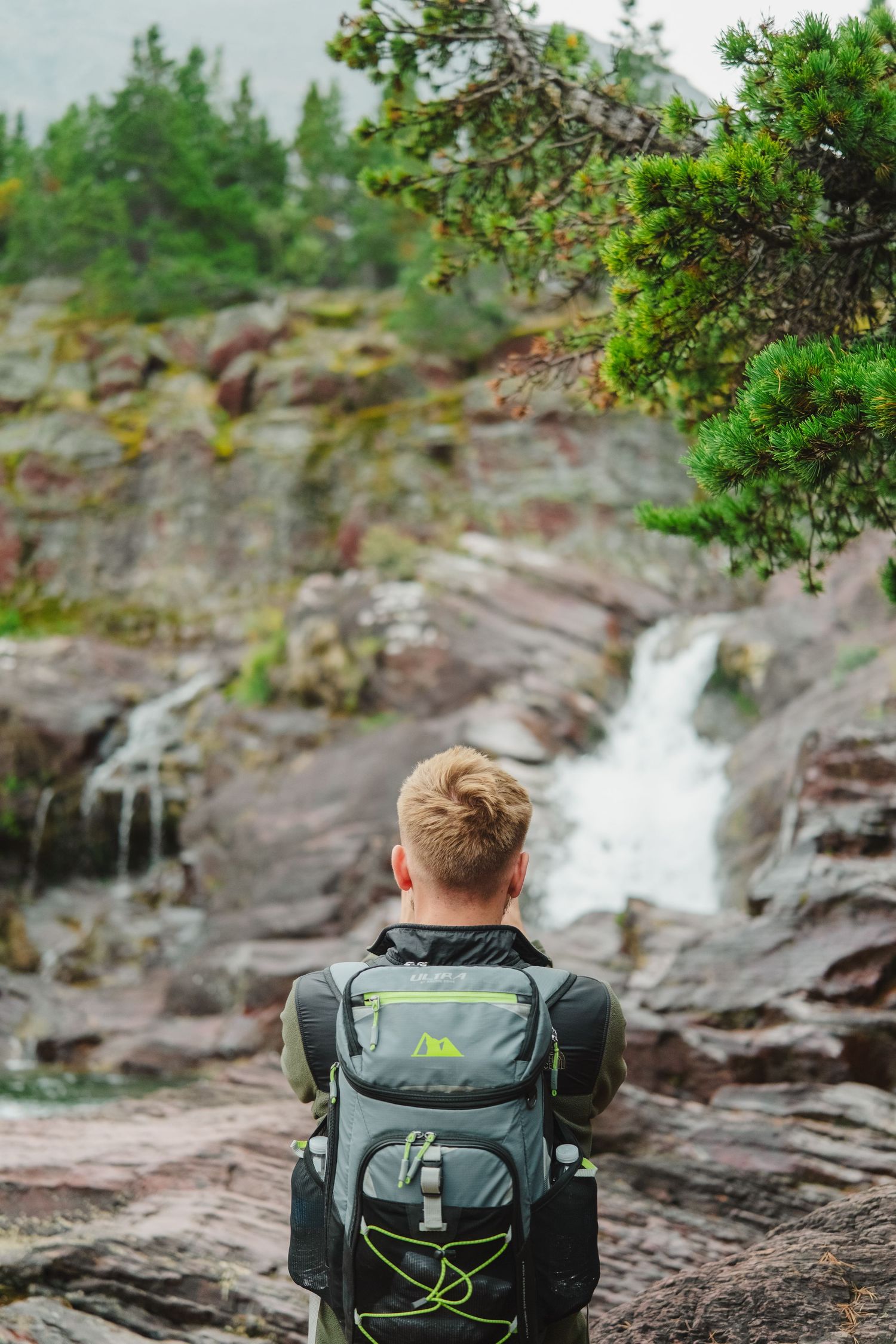 Backpack sitting on rocky cliff edge overlooking scenic waterfall in misty forest landscape.