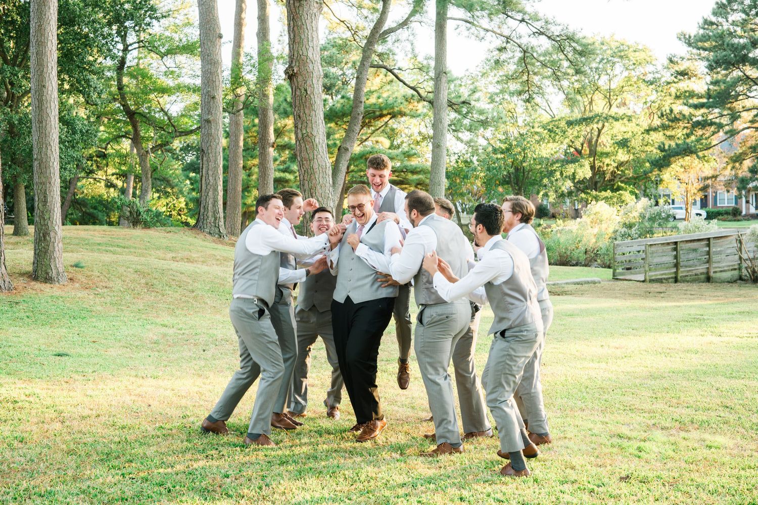 Wedding party in gray suits playfully pose together on a grassy lawn surrounded by trees on a sunny day.