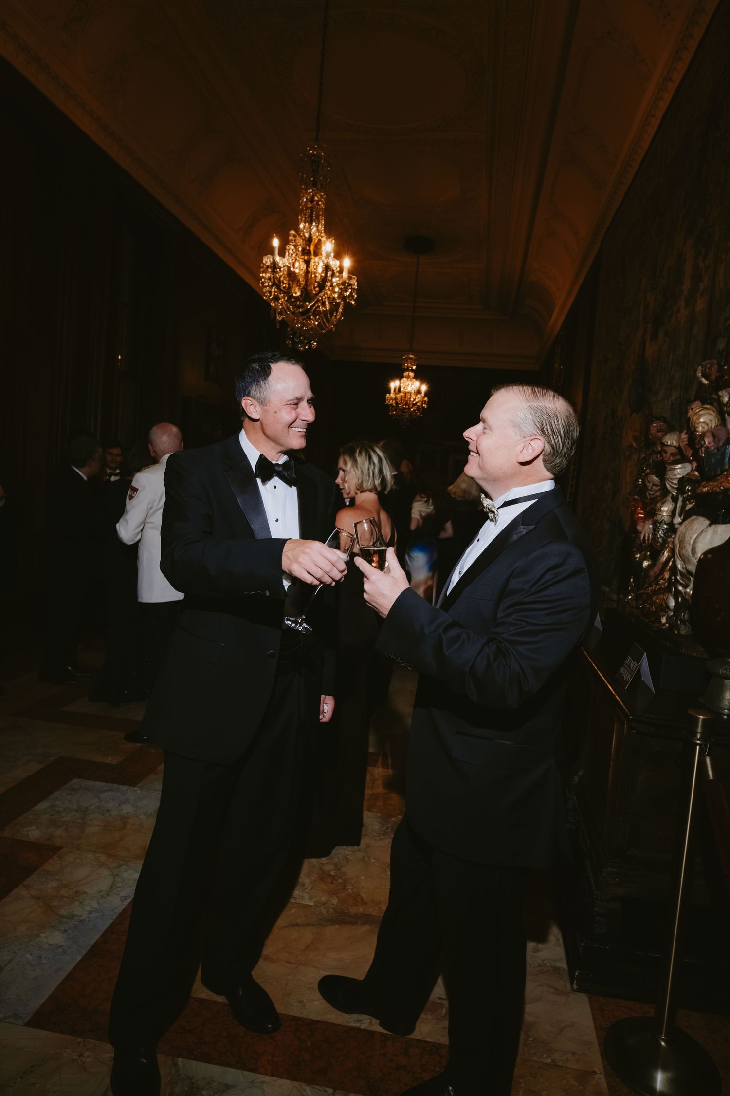Two men in tuxedos dancing and laughing together at an elegant event with chandeliers in the background.