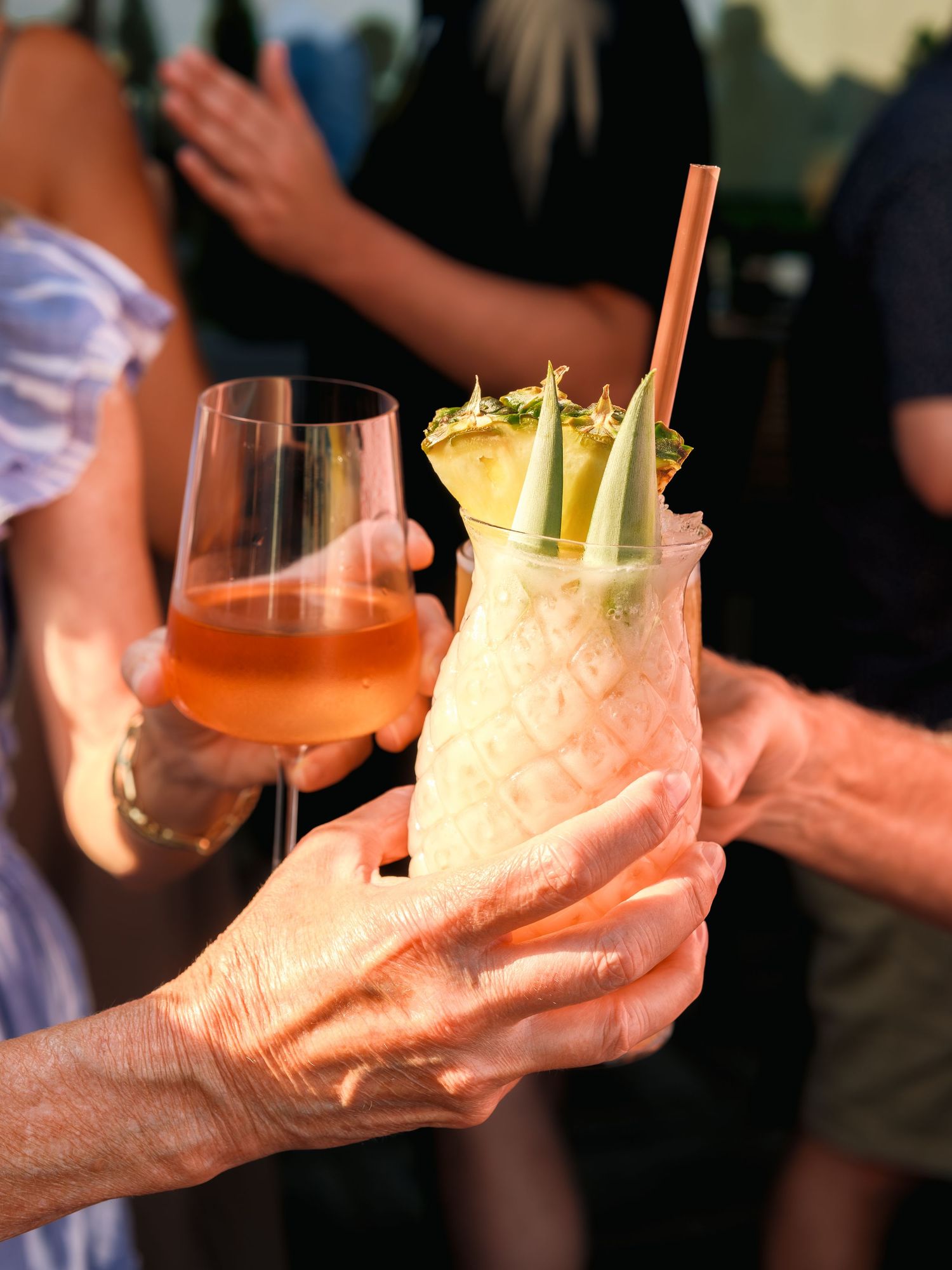 Two hands hold cocktail drinks, one with pink liquid and garnishes and another with an orange-colored beverage.