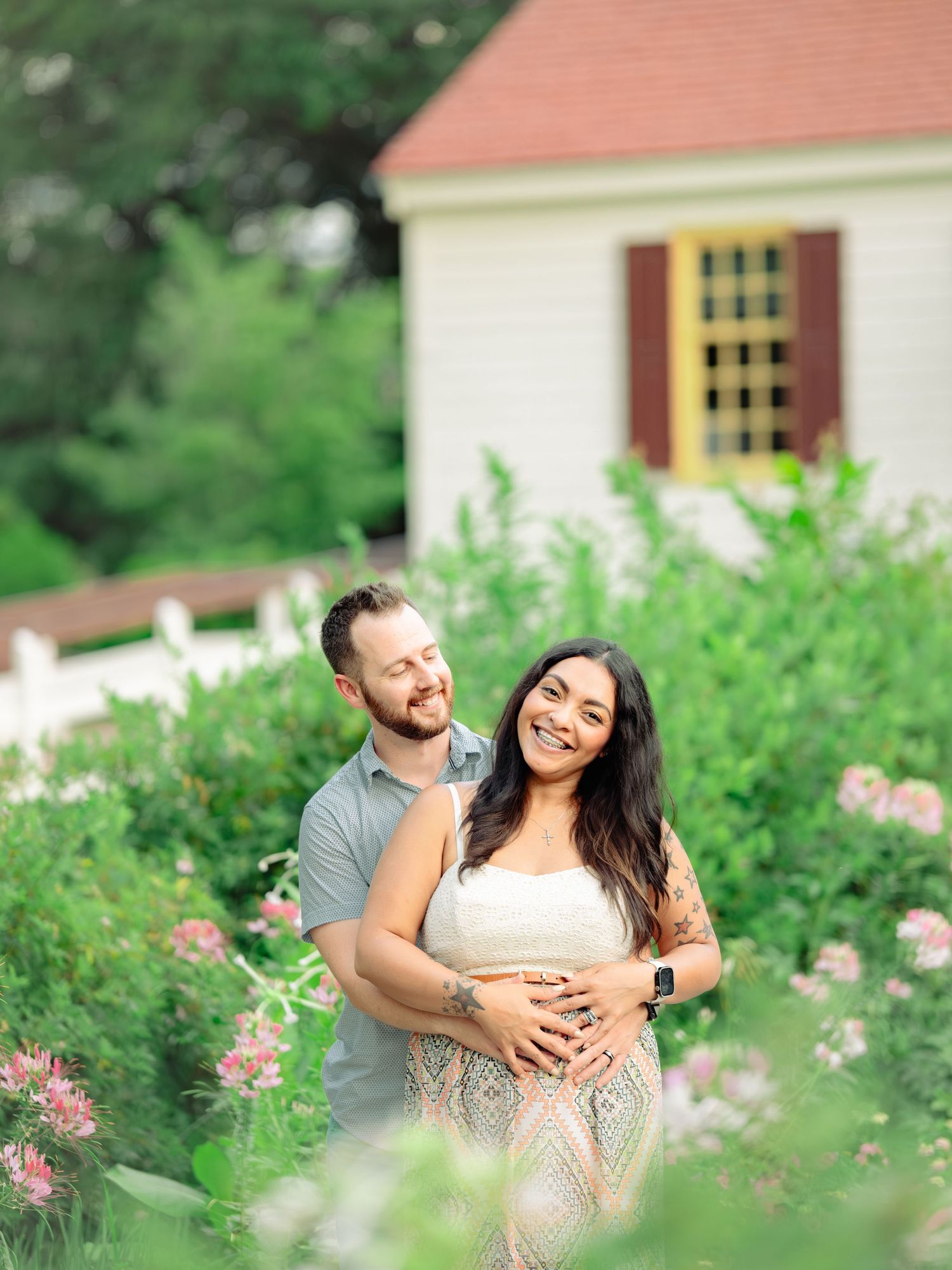 A loving couple shares a tender moment among pink flowers and greenery outside a house with a red roof.
