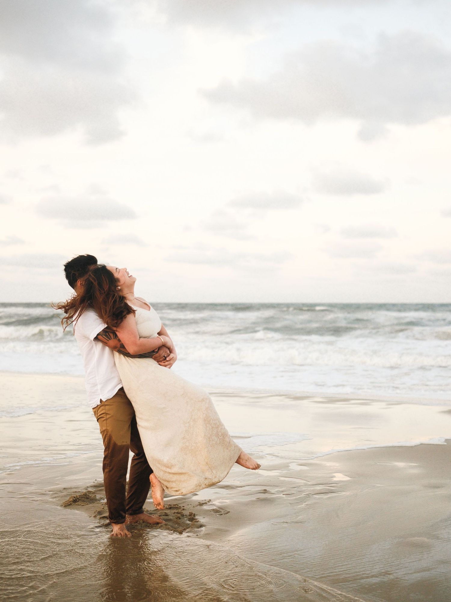 Couple playing in ocean waves at sunset while bride's dress gets wet.