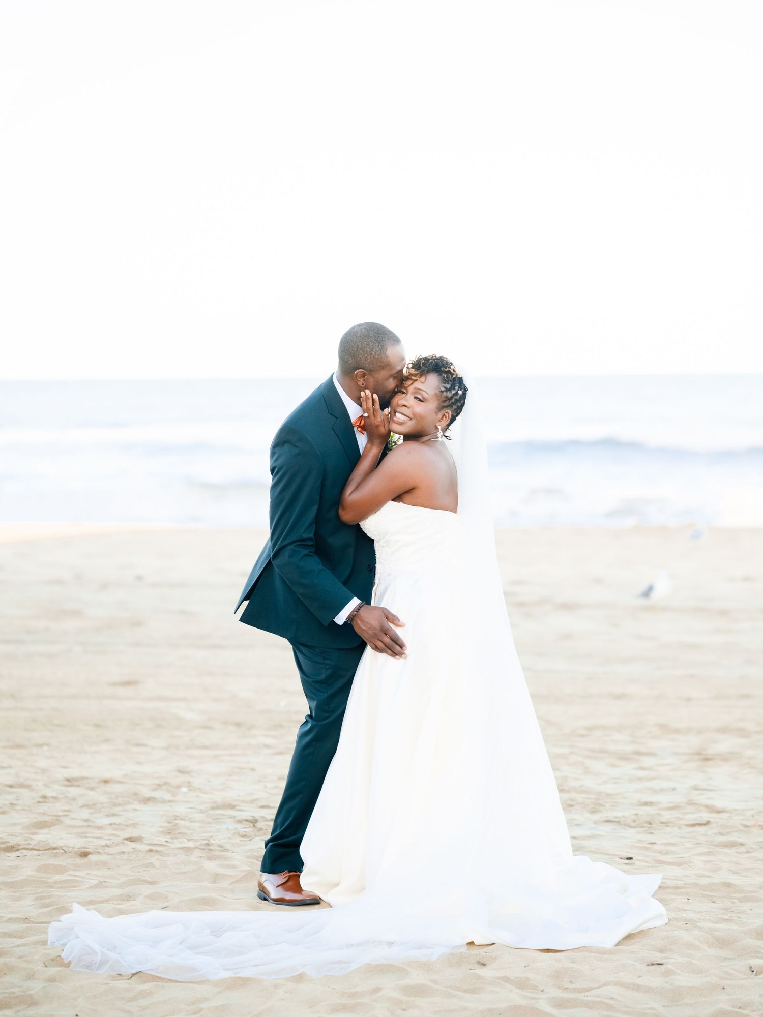A romantic beach wedding scene shows a couple in formal attire sharing a tender moment on the sand at sunset.