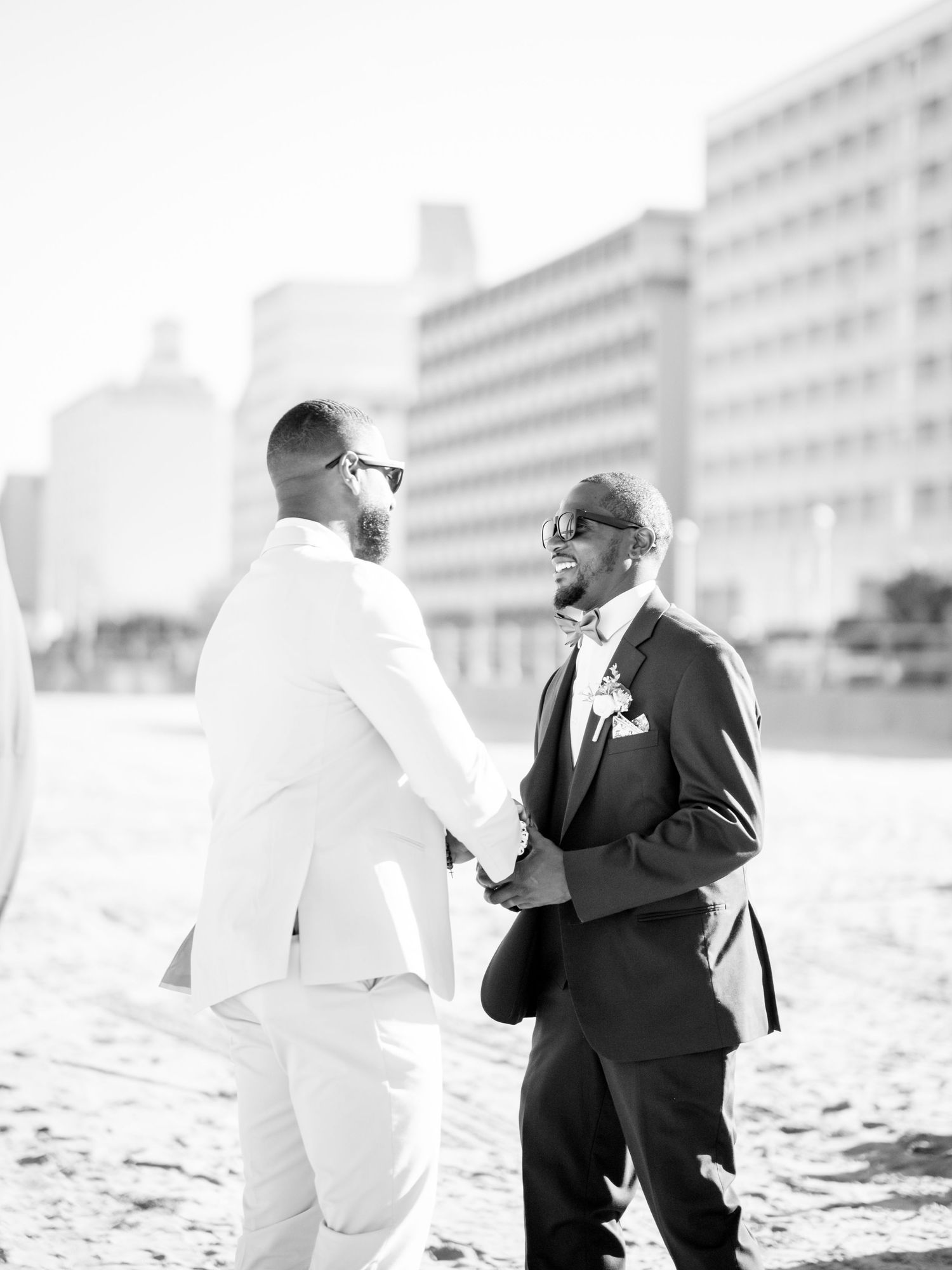 Two people in contrasting white and black suits sharing a joyful moment on a beach with buildings in background.