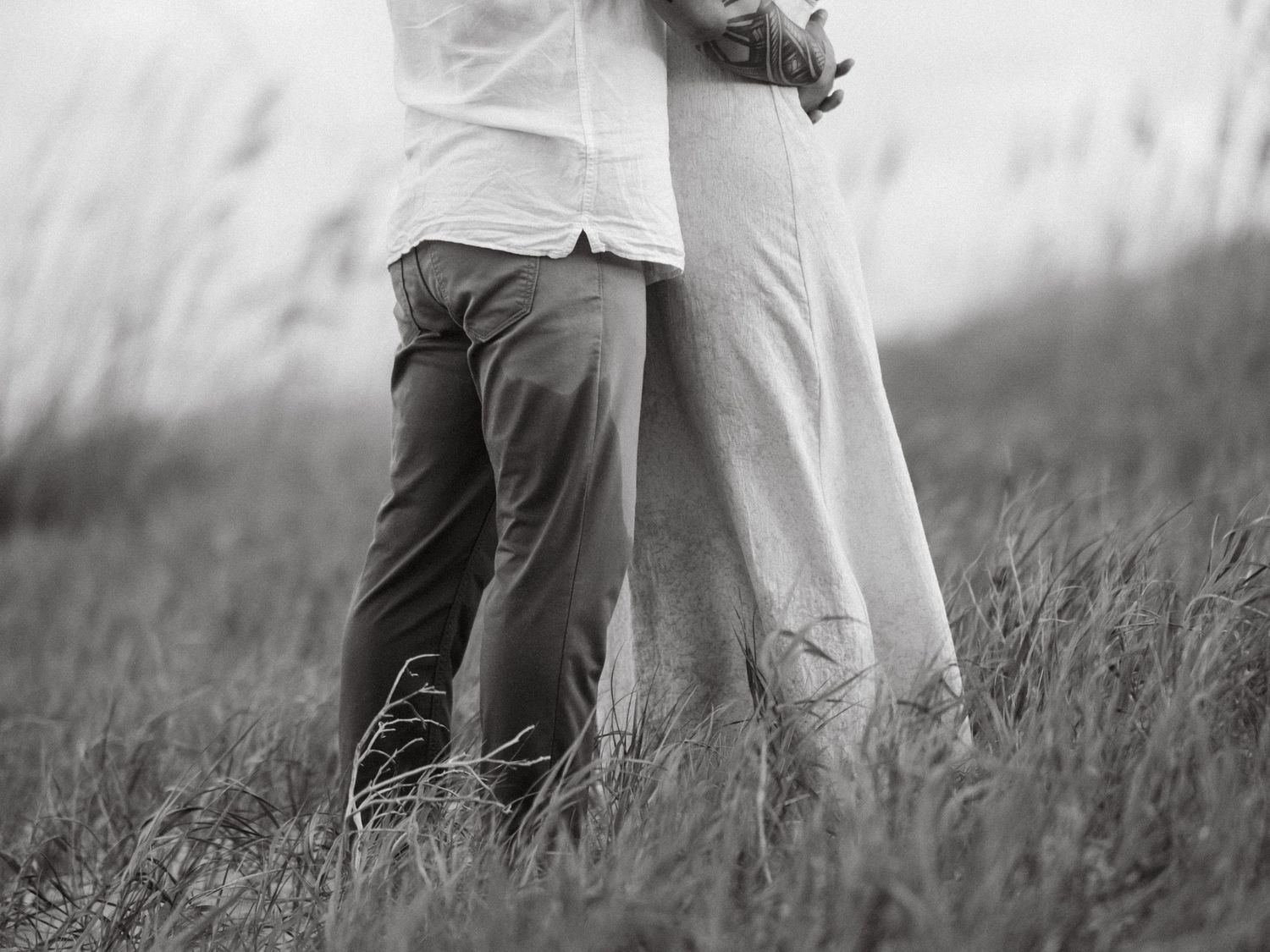 Black and white field engagement photo of two people holding hands and walking through tall grass at sunset.