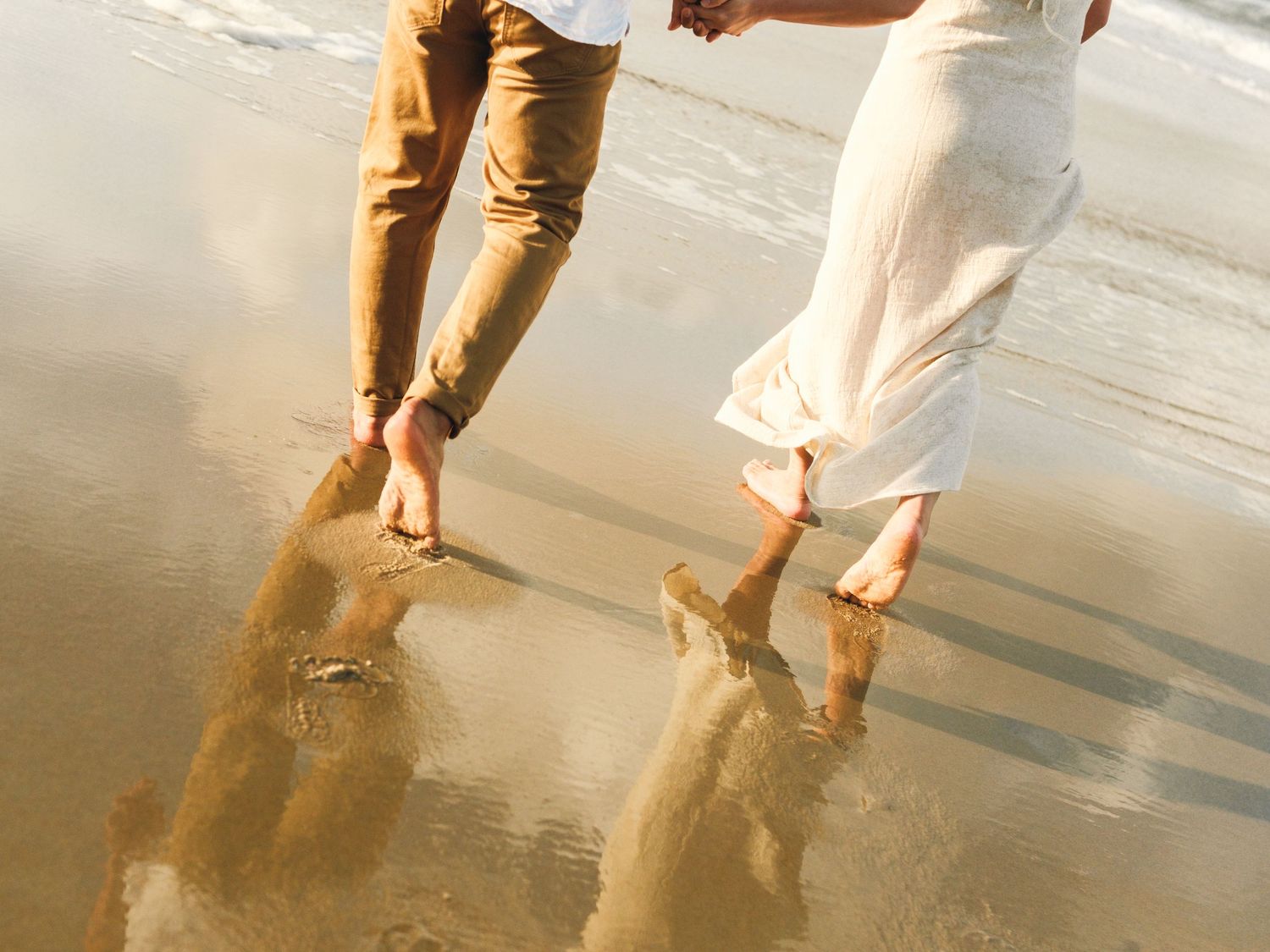 Couple walking barefoot together along a sandy beach shoreline at sunset.
