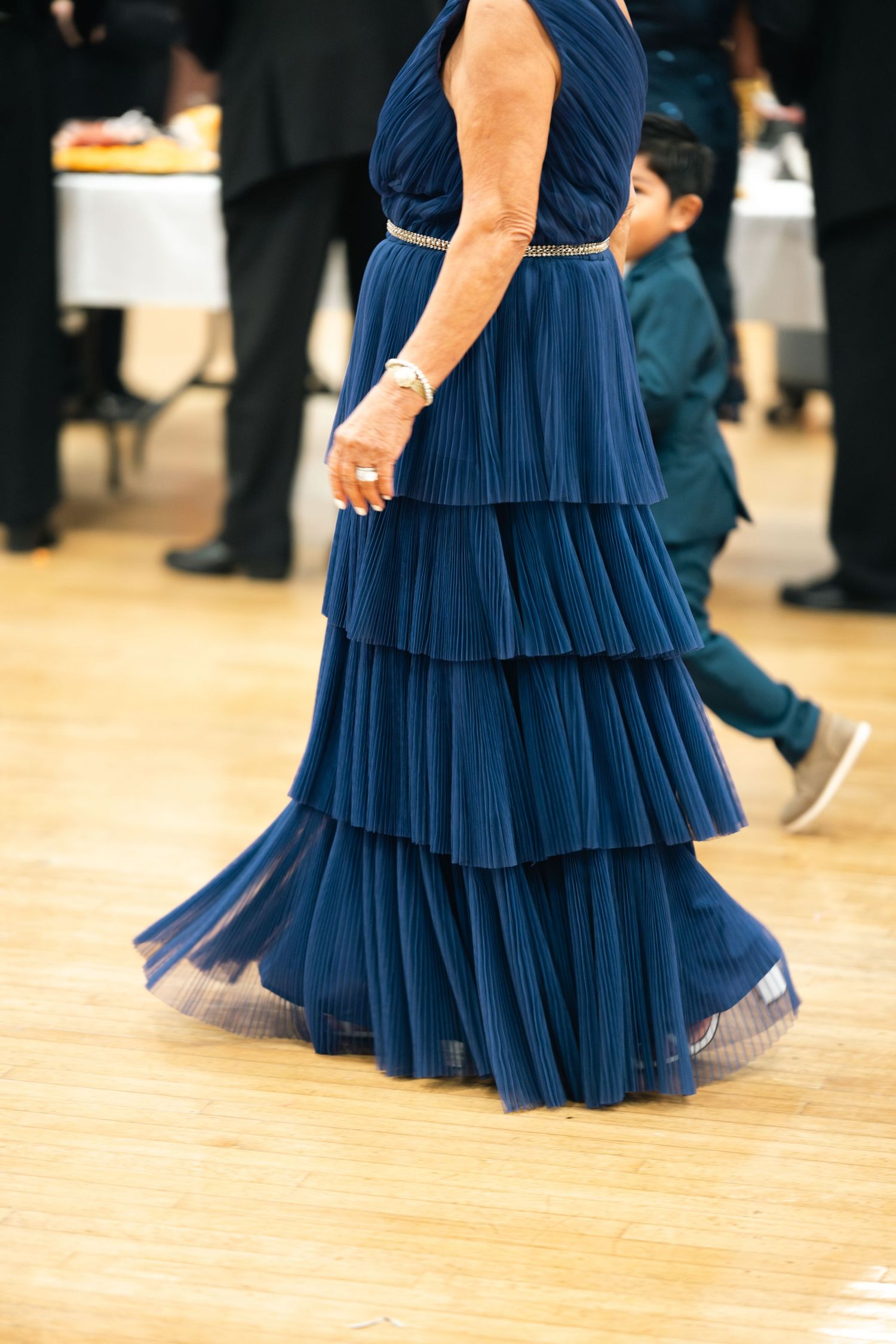 Woman wearing an elegant navy blue tiered pleated maxi dress stands on wooden dance floor at formal event.