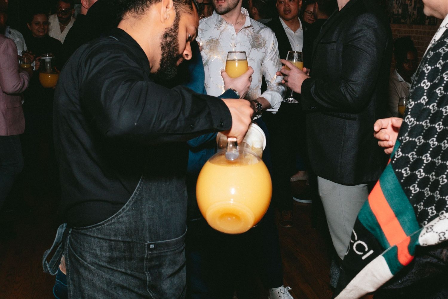 People at a party gather around while someone pours a drink into a large yellow balloon during a social gathering.