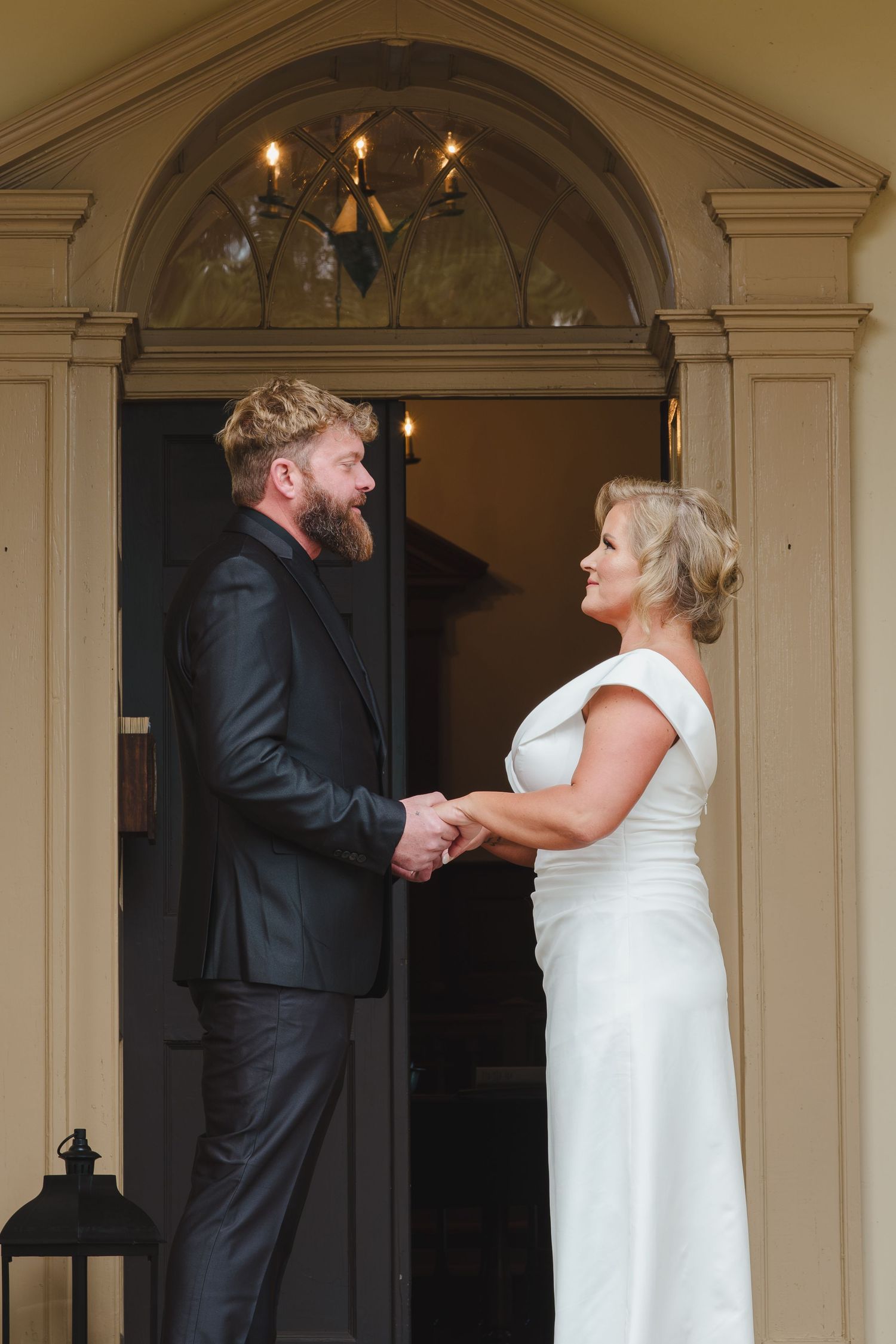 A couple shares a tender moment holding hands in formal attire at an elegant entryway with arched doorway and lantern.
