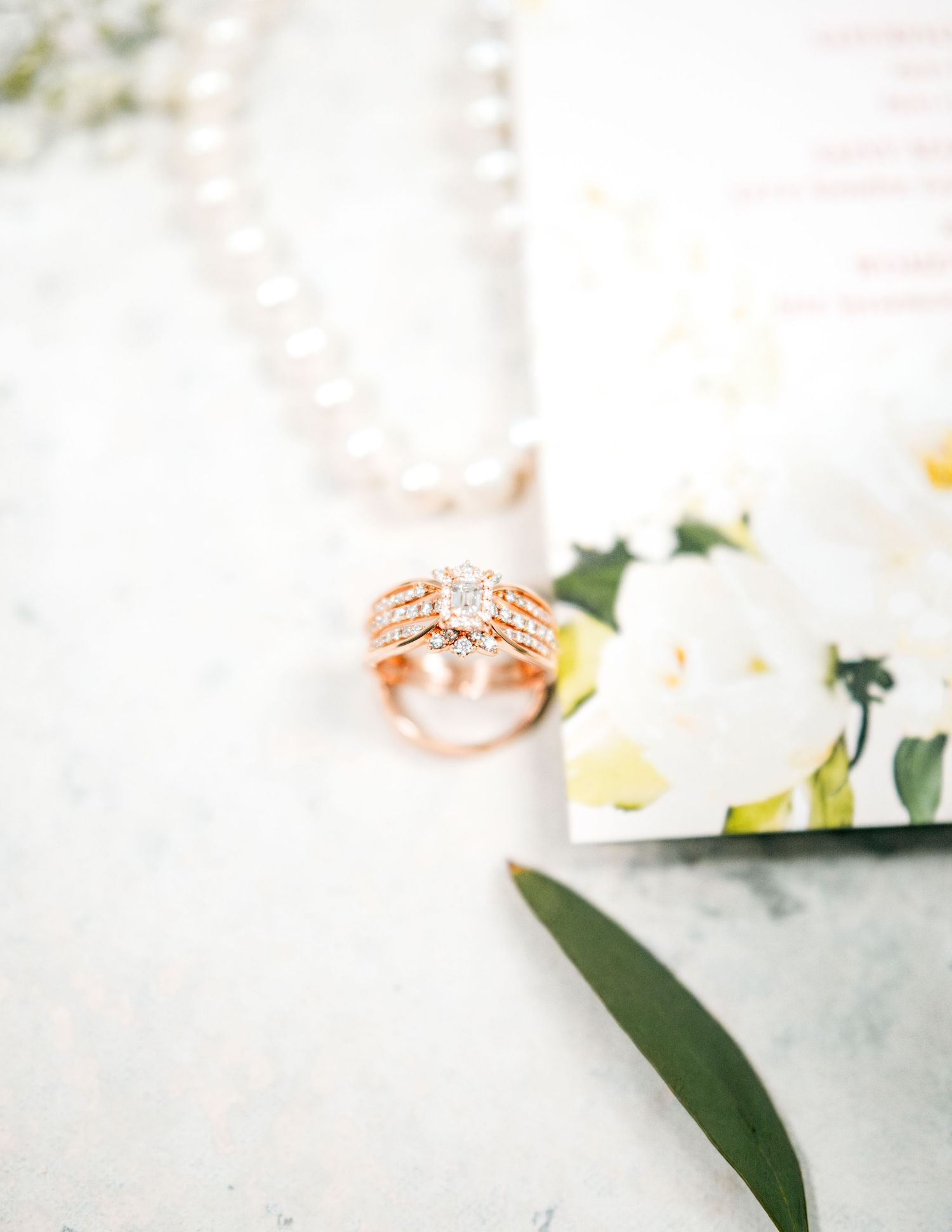 Rose gold engagement ring with diamonds sits near white flowers and green foliage on white marble surface.