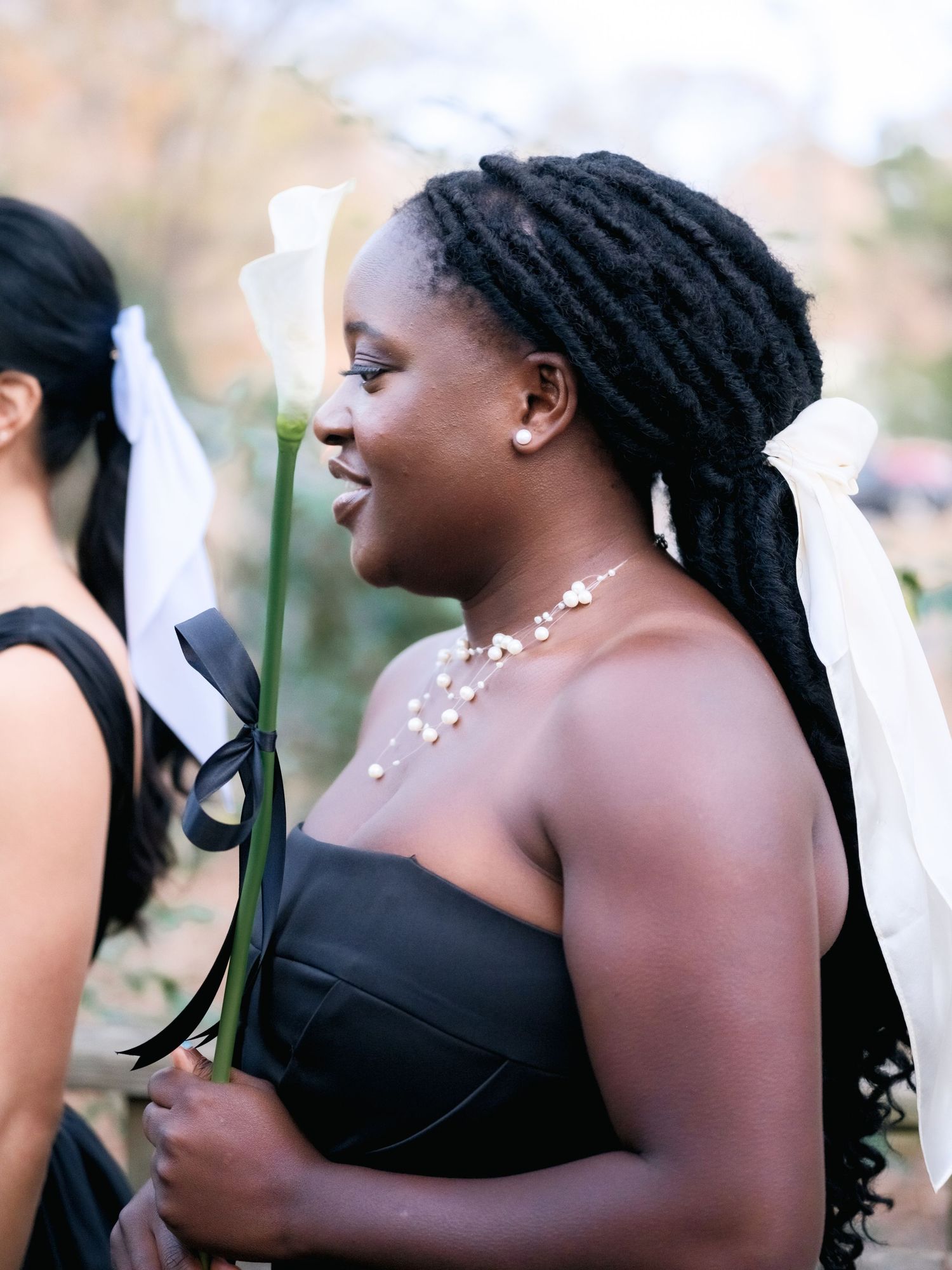 Bride in a black strapless dress holds a white calla lily at a daytime outdoor wedding ceremony.