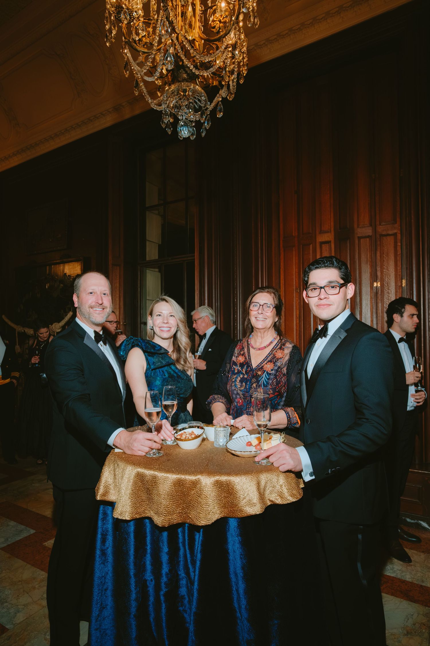 Wedding guests socialize at elegant round tables decorated with navy blue linens and crystal chandeliers overhead.