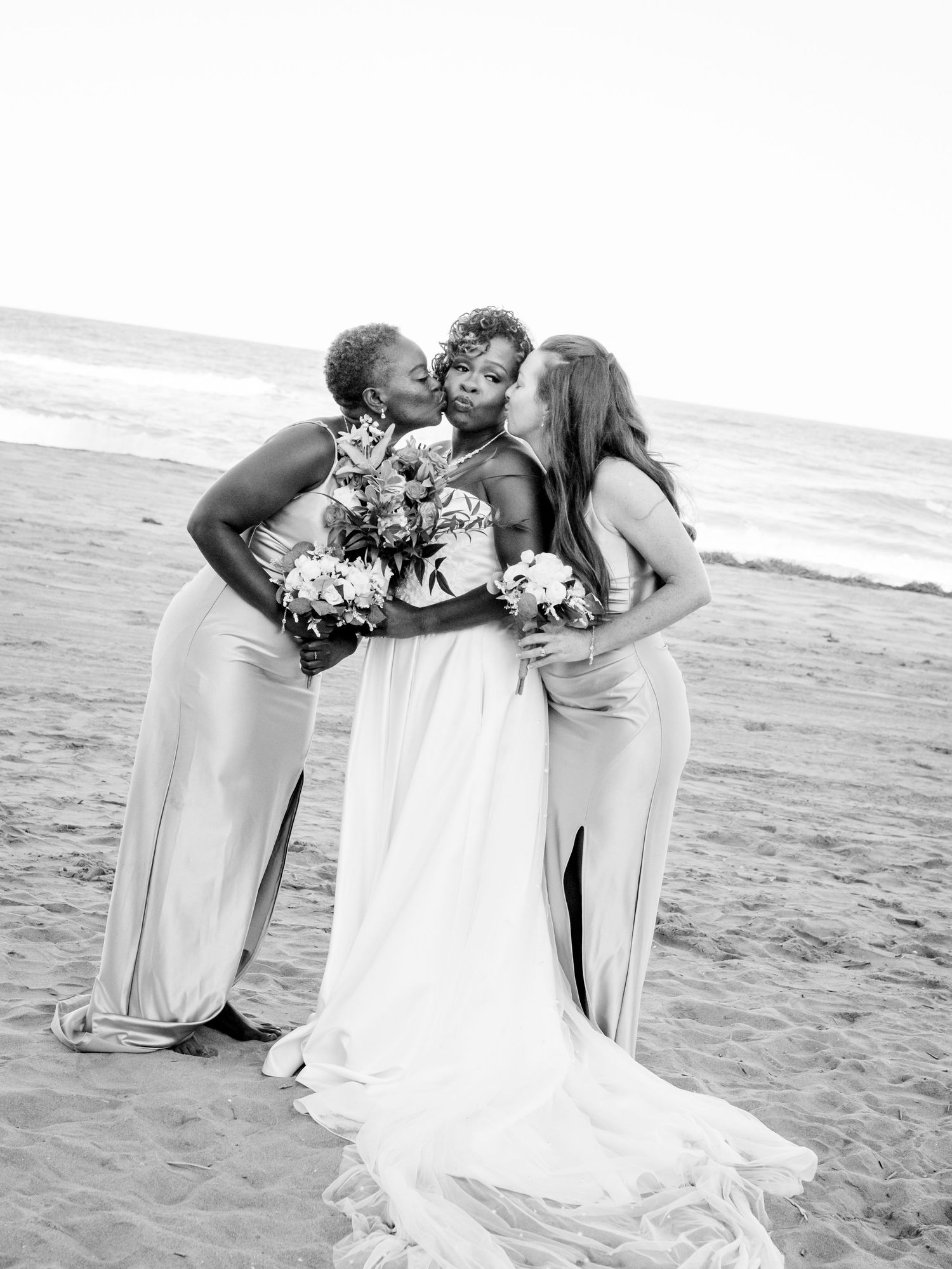 Three people in white dresses embrace on a beach during a wedding celebration.