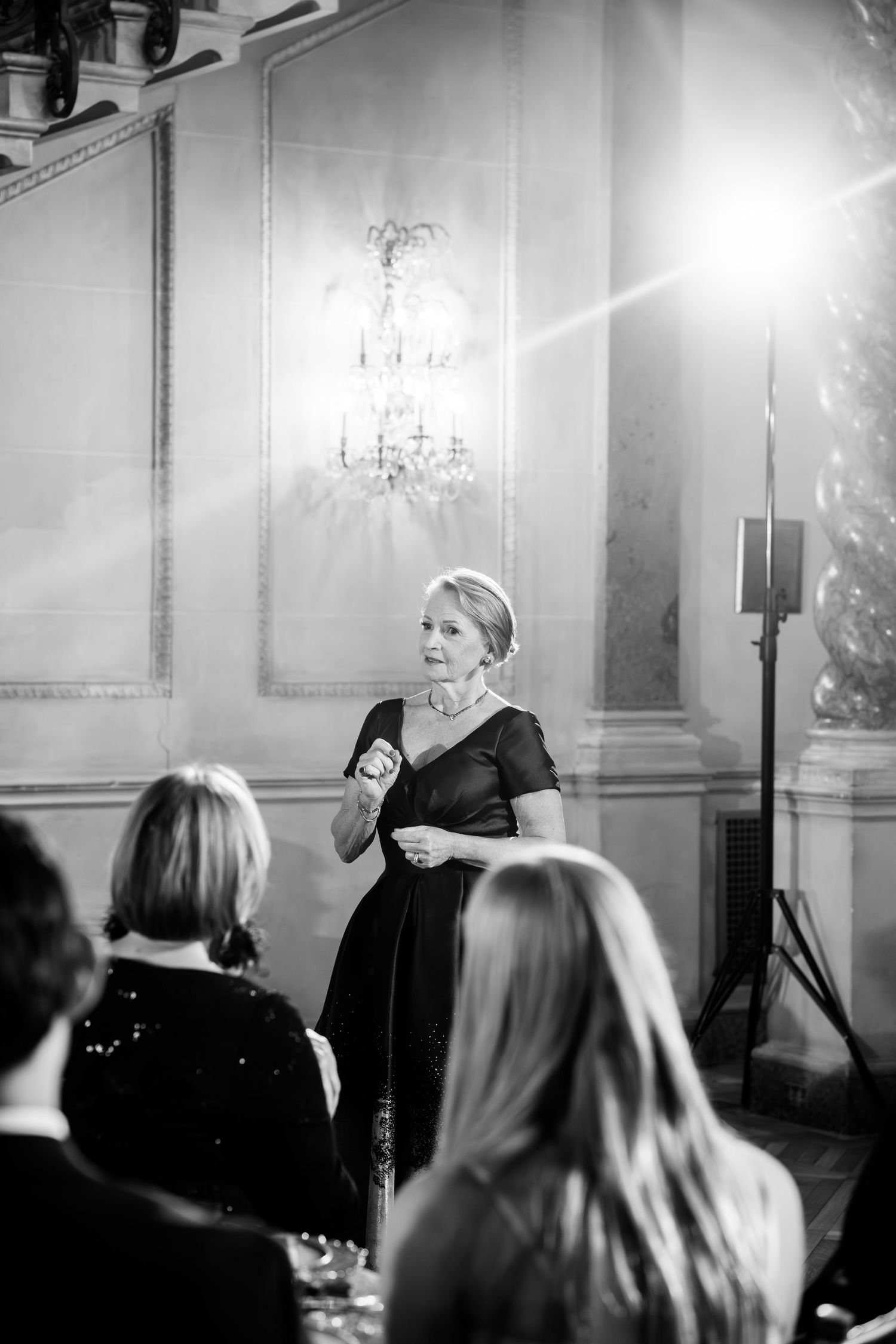 A performer sings on stage in a dimly lit venue with elegant chandeliers and an attentive audience in silhouette.