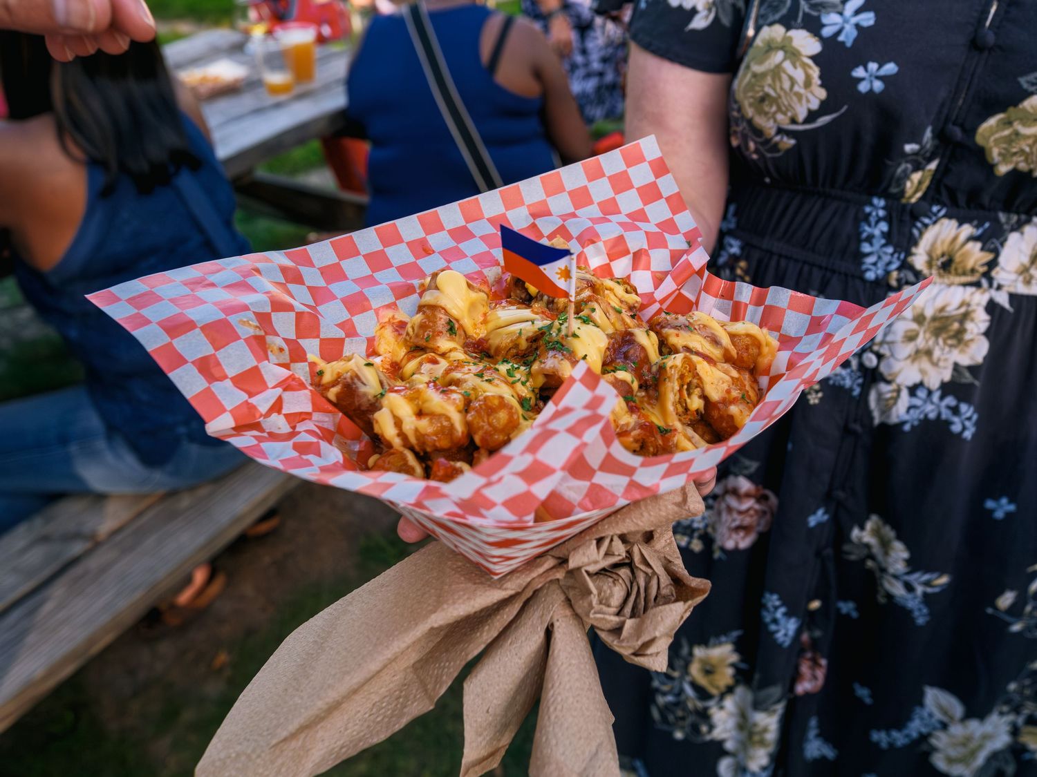 Two hands hold fresh garlic knots and chicken wings served in traditional red and white checkered paper boats.
