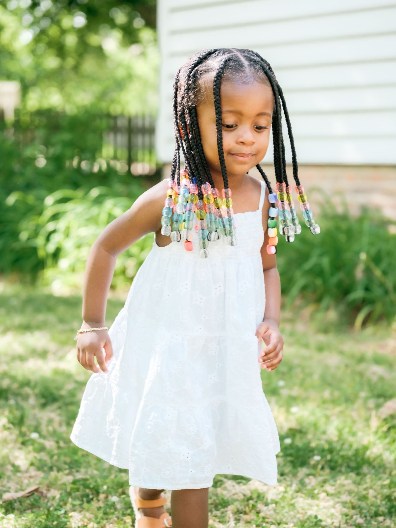 A young child in a white summer dress twirls playfully outdoors, wearing colorful beaded braids in their hair.