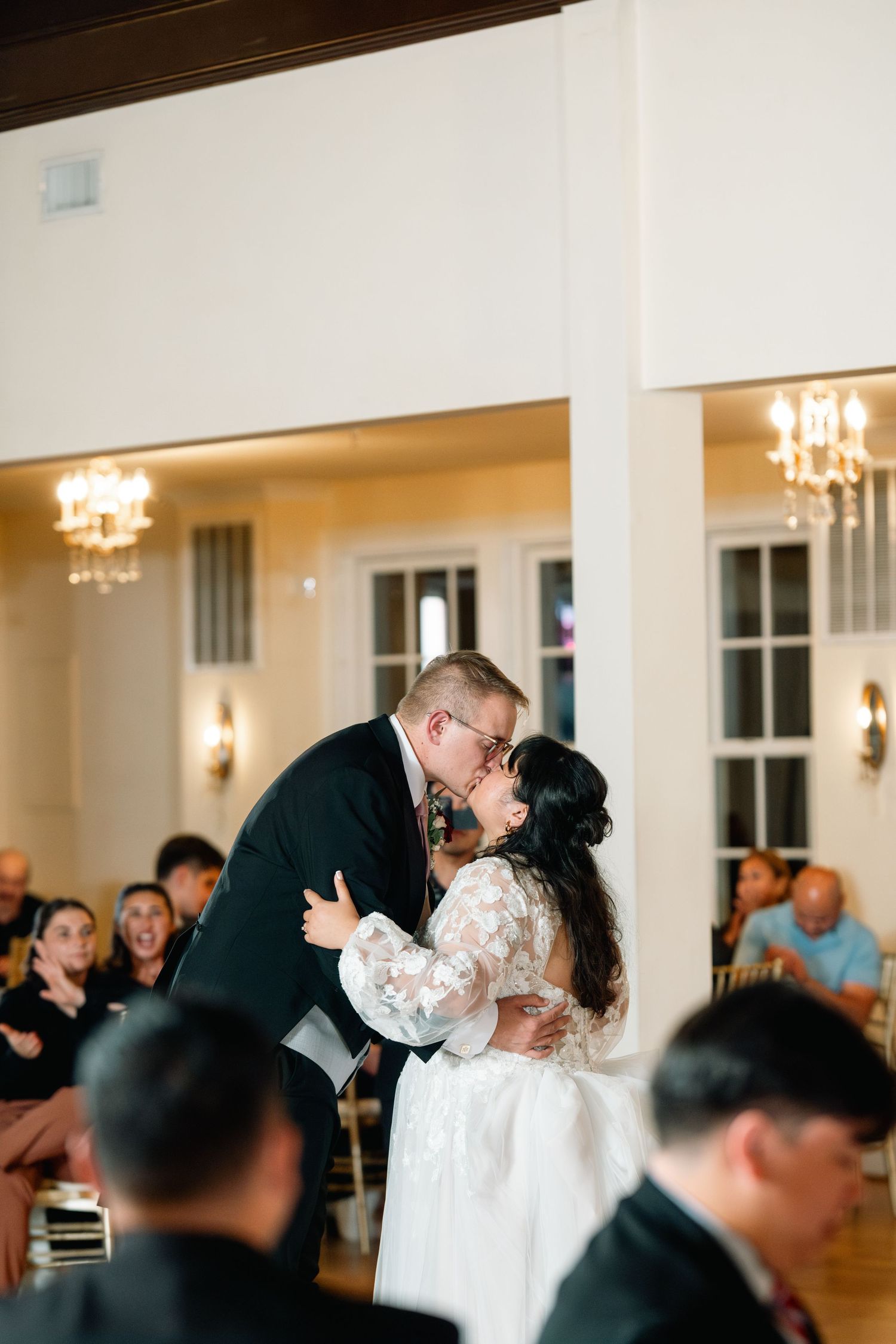Couple shares their first dance in an elegant ballroom with chandeliers and watching guests at their wedding reception.