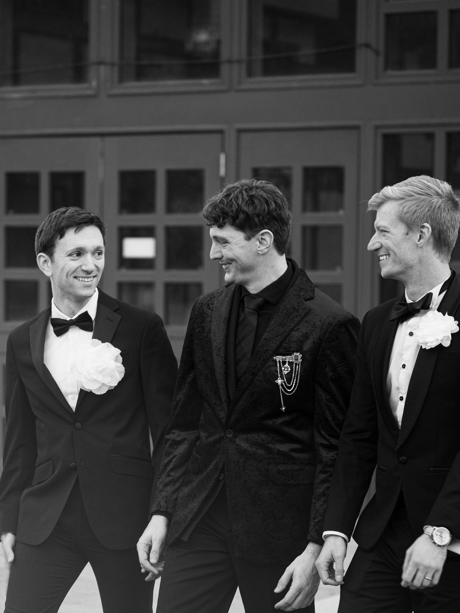 Black and white candid photo of groomsmen laughing together in formal tuxedos at a wedding celebration.