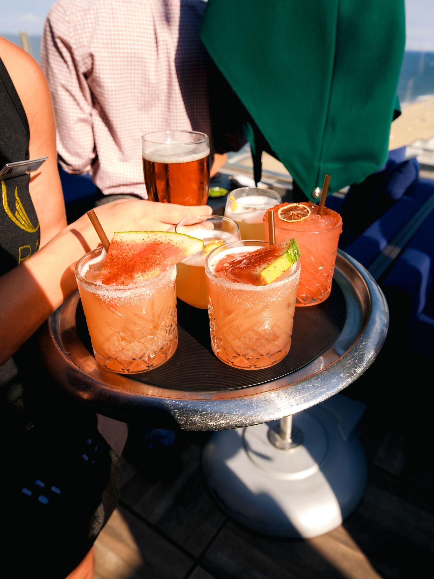A tray of pink cocktails garnished with citrus slices and a beer glass in the background on a sunny day.
