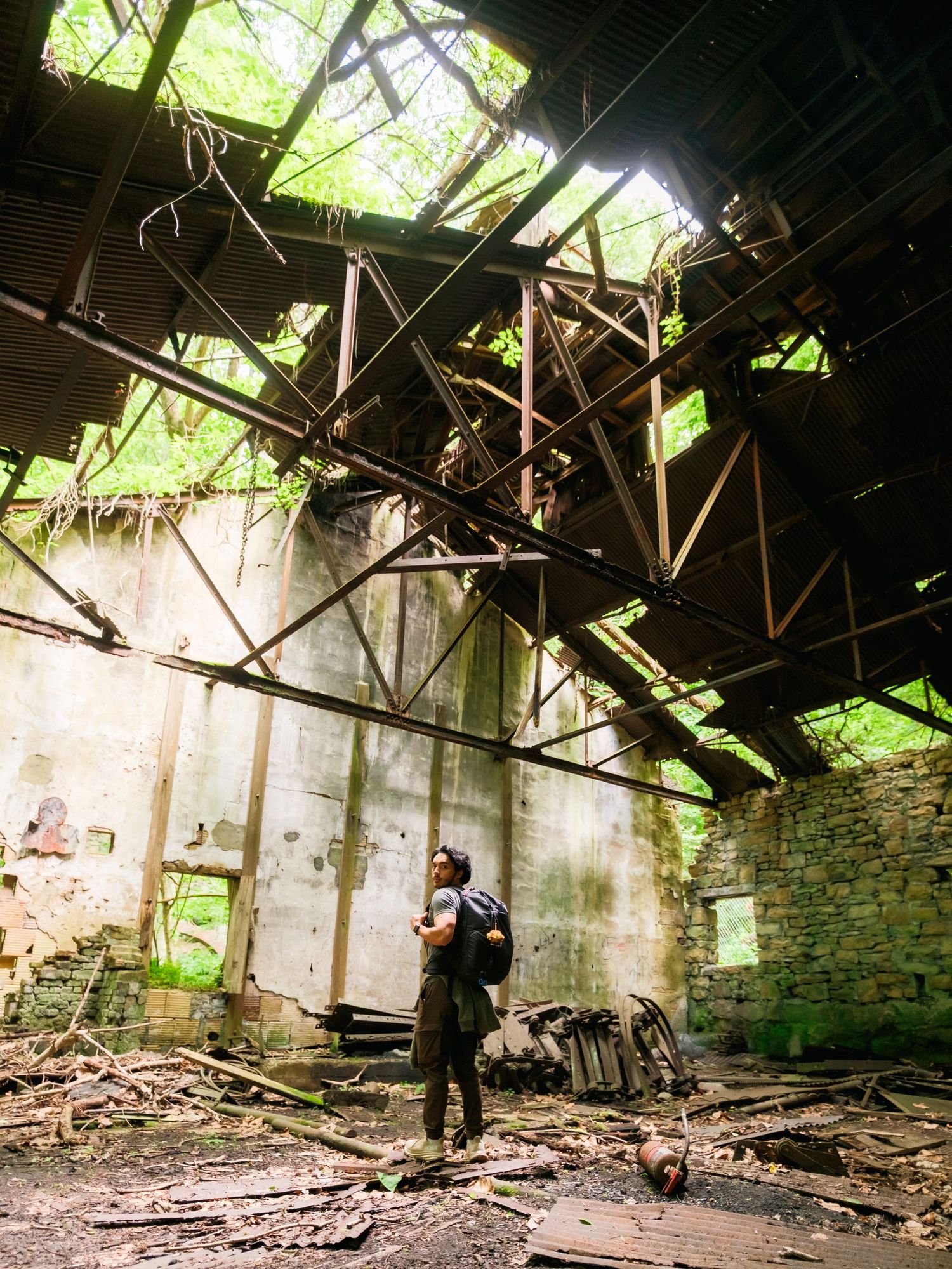 Photographer exploring abandoned building with broken ceiling and green foliage growing through exposed rafters above.
