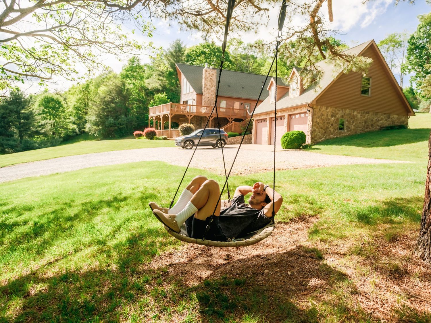 A tire swing hangs in front of a rustic log cabin on a sunny grassy hill with a brown outbuilding nearby.