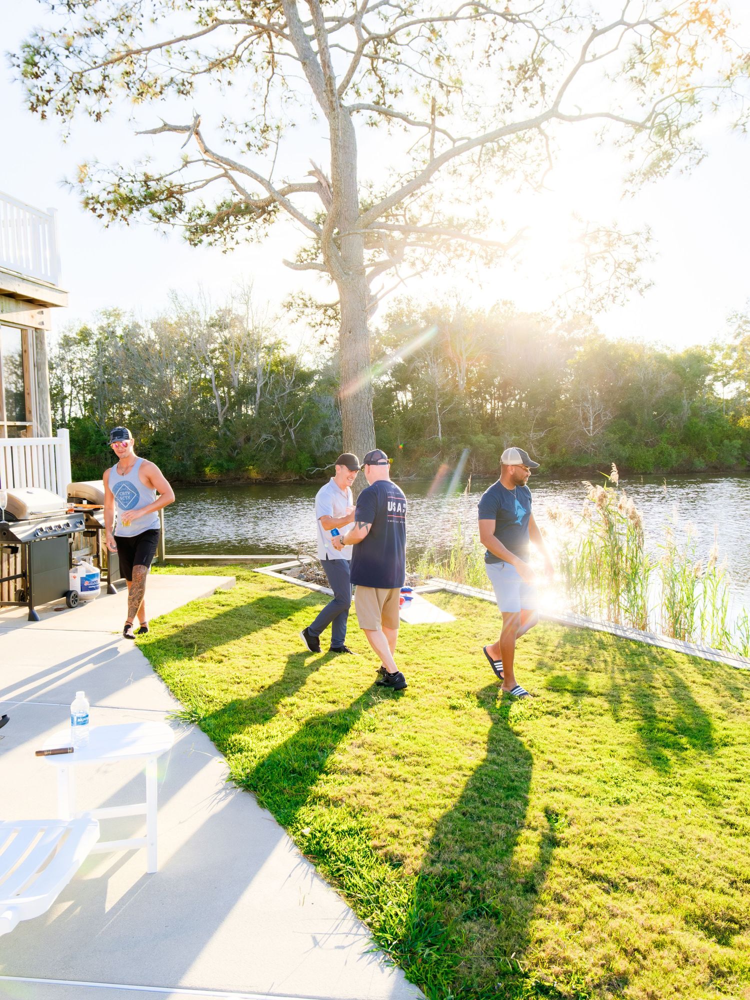 A group of people enjoying a sunny afternoon gathering on a waterfront lawn with golden sunlight shining through trees.
