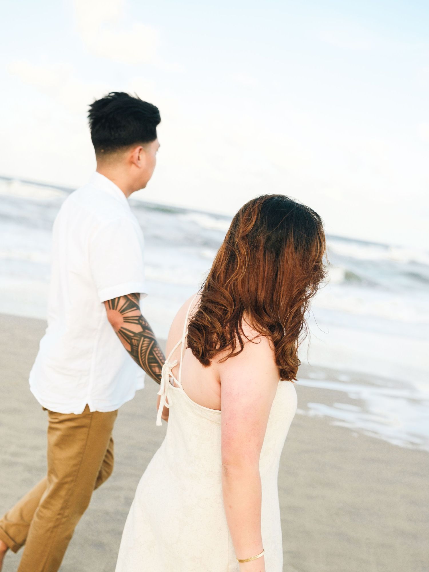 Romantic couple taking a stroll on sandy beach at sunset wearing white and brown casual attire.
