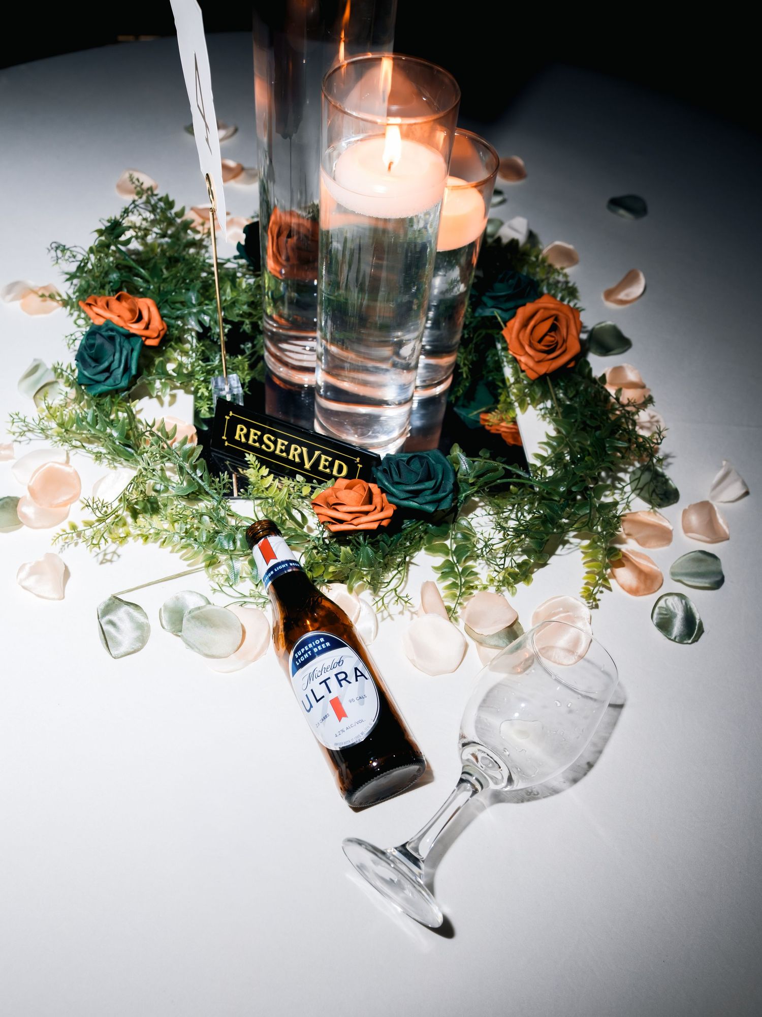 Wedding reception table decor with orange roses, greenery, candles, and scattered liquor bottles.