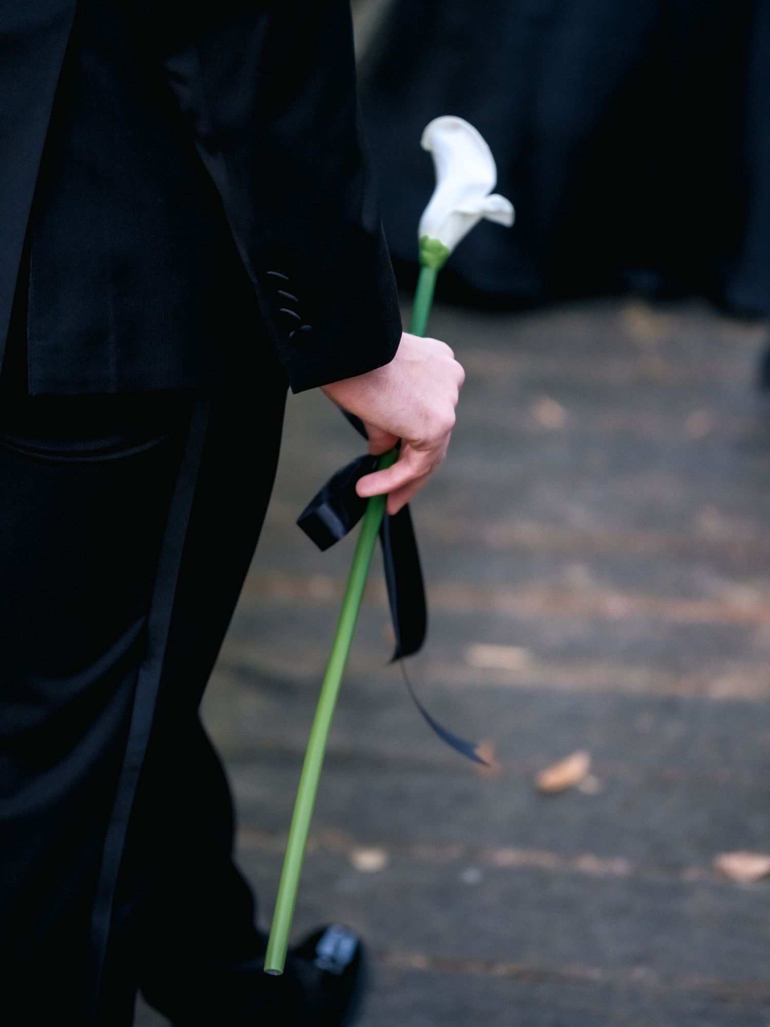 A single white calla lily stem with long green stalk is held against a dark background by someone wearing formal attire.