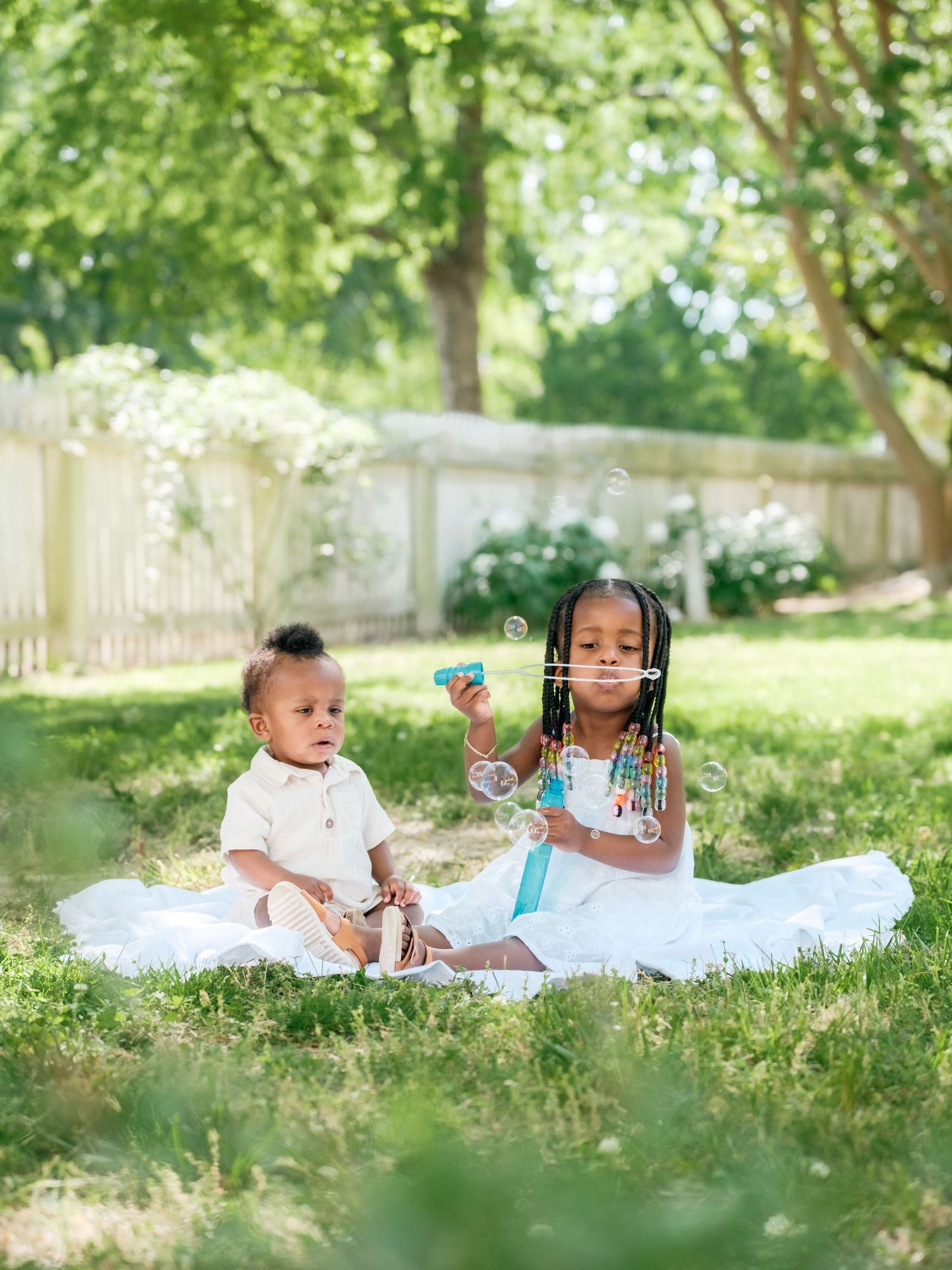 Two children sitting on a white blanket in a sunny backyard with flowers and a white fence in the background.