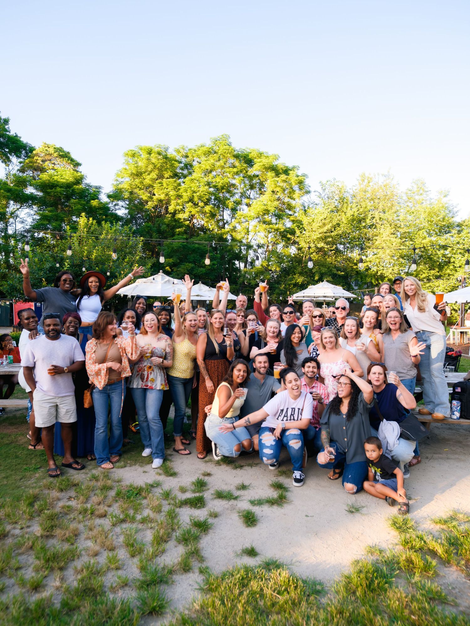 A large group poses together outdoors on a sunny day, holding white parasols and drinks while celebrating.