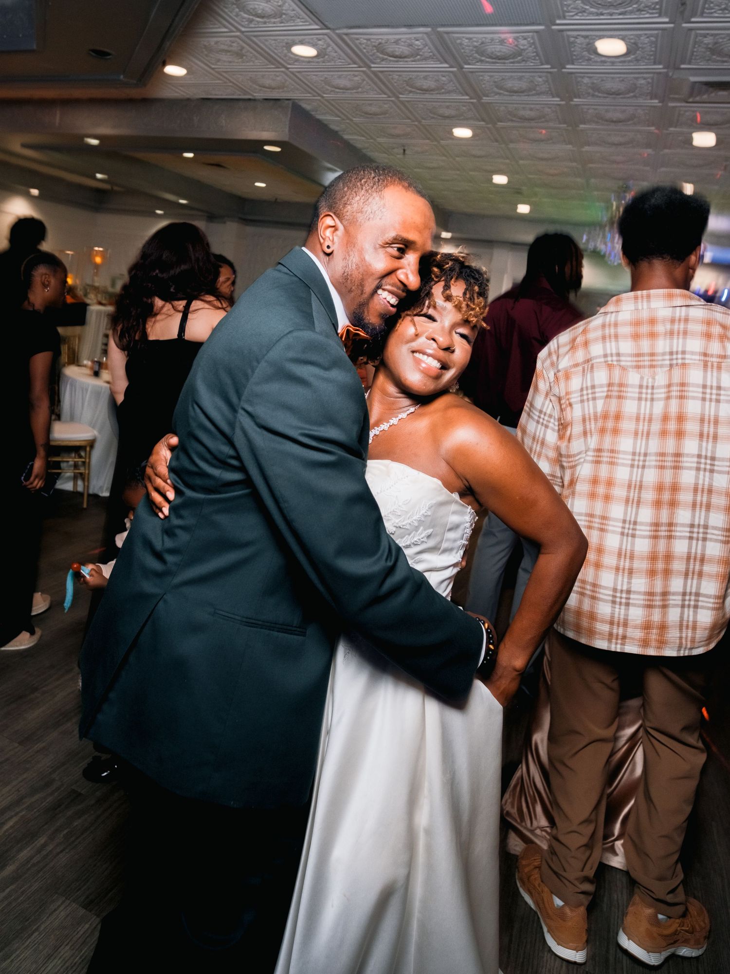 A couple shares an intimate dance moment at a reception venue with other guests visible in the background.