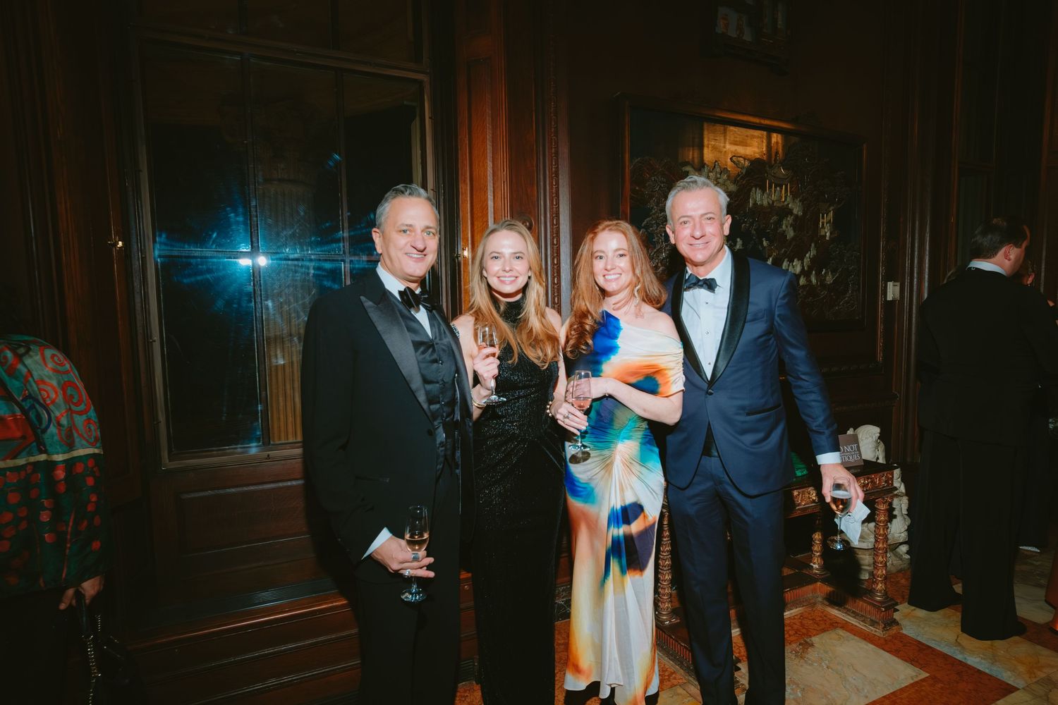 Four people in formal evening attire pose together at an elegant indoor event with dark wood paneled walls.