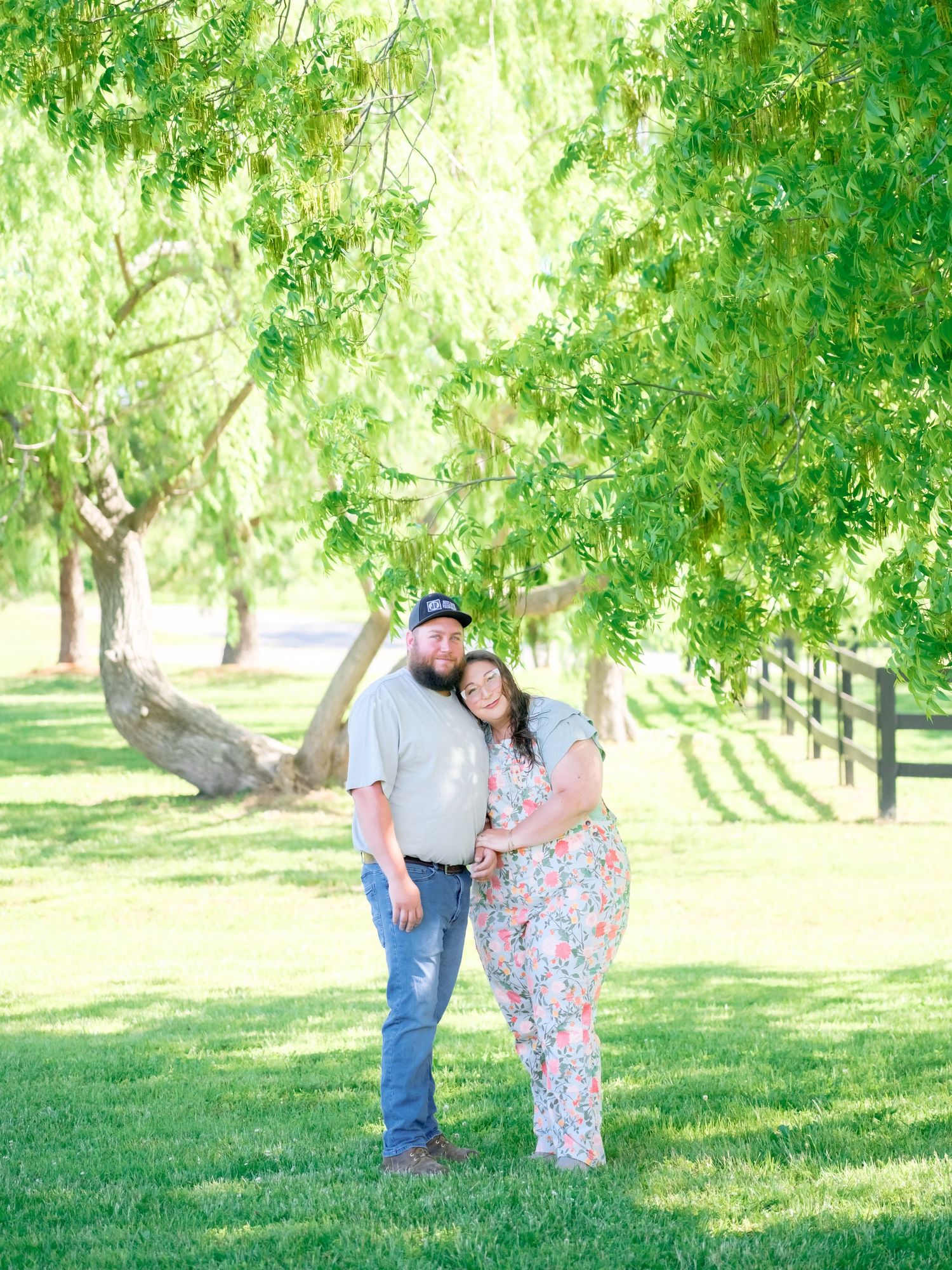 A couple stands together in a romantic outdoor setting under green trees on a sunny day.
