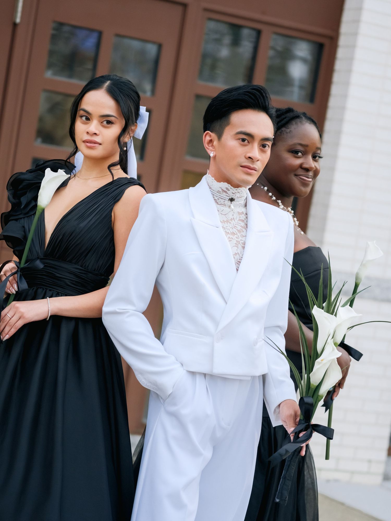Three people dressed in formal attire stand together, with one in a black gown and another in an elegant white suit.