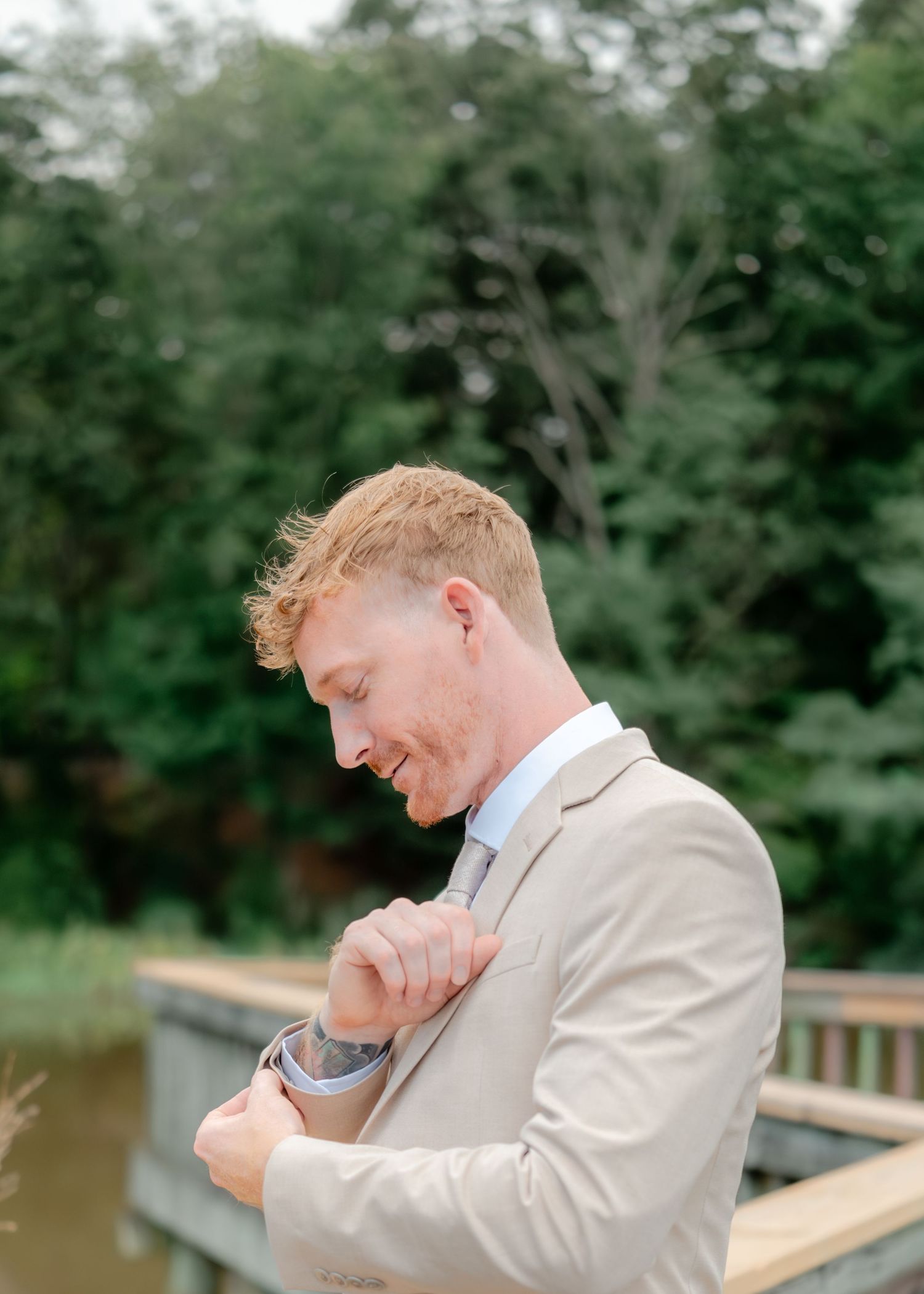 A person wearing a light beige suit adjusts their sleeve while standing on a wooden deck near water and trees.