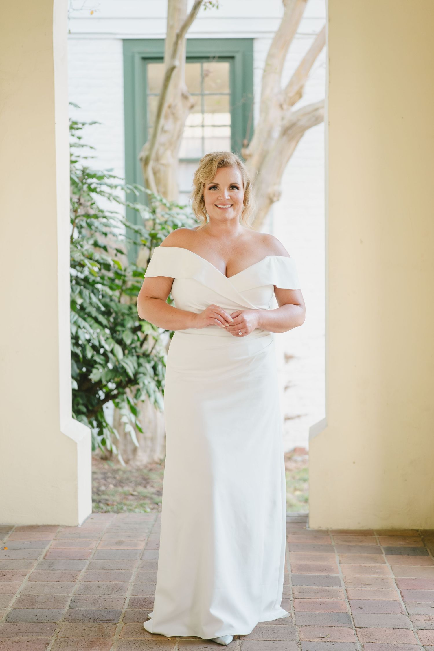 An elegant off-shoulder white wedding gown is worn on a tiled patio with a mint green door frame and greenery in view.